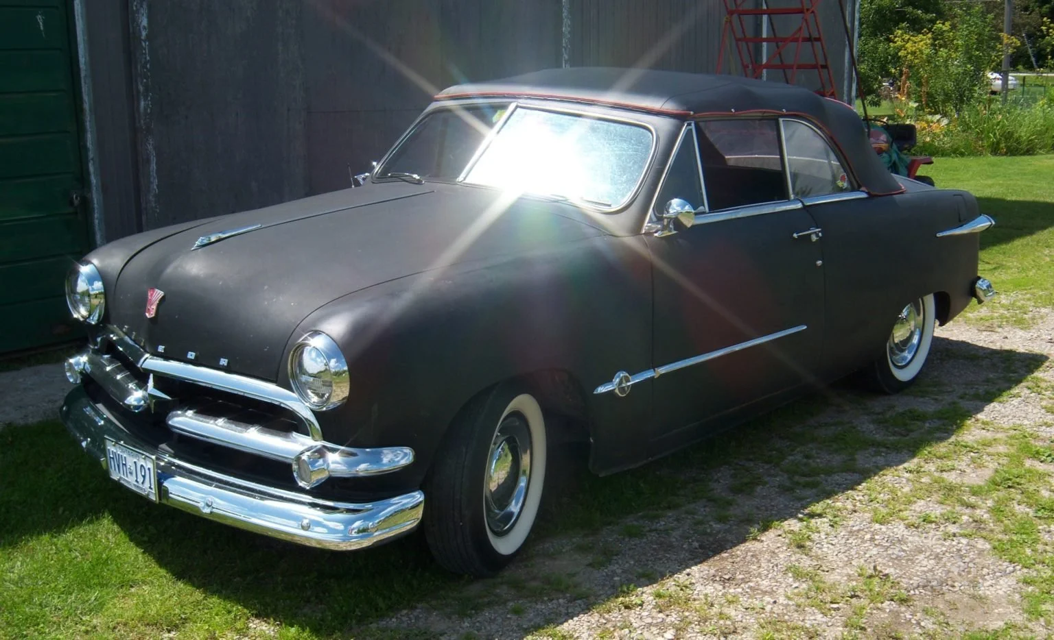 A vintage black convertible car parked on a grassy area beside a gravel path, with chrome details, whitewall tires, and a soft top roof, sunlight reflecting off its surface.
