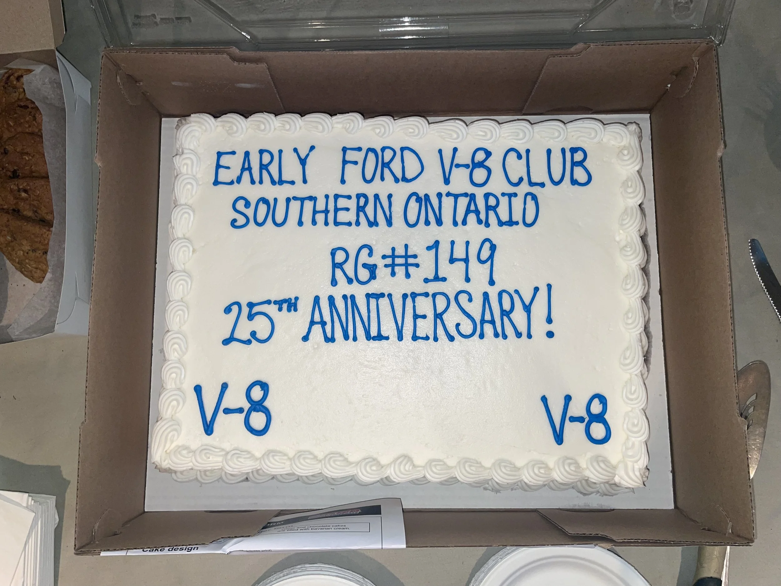 Celebration cake with white frosting and blue writing that reads 'Early Ford V-8 Club, Southern Ontario, RG#149, 25th Anniversary!' and the initials V-8 on the bottom corners, inside a cardboard box.