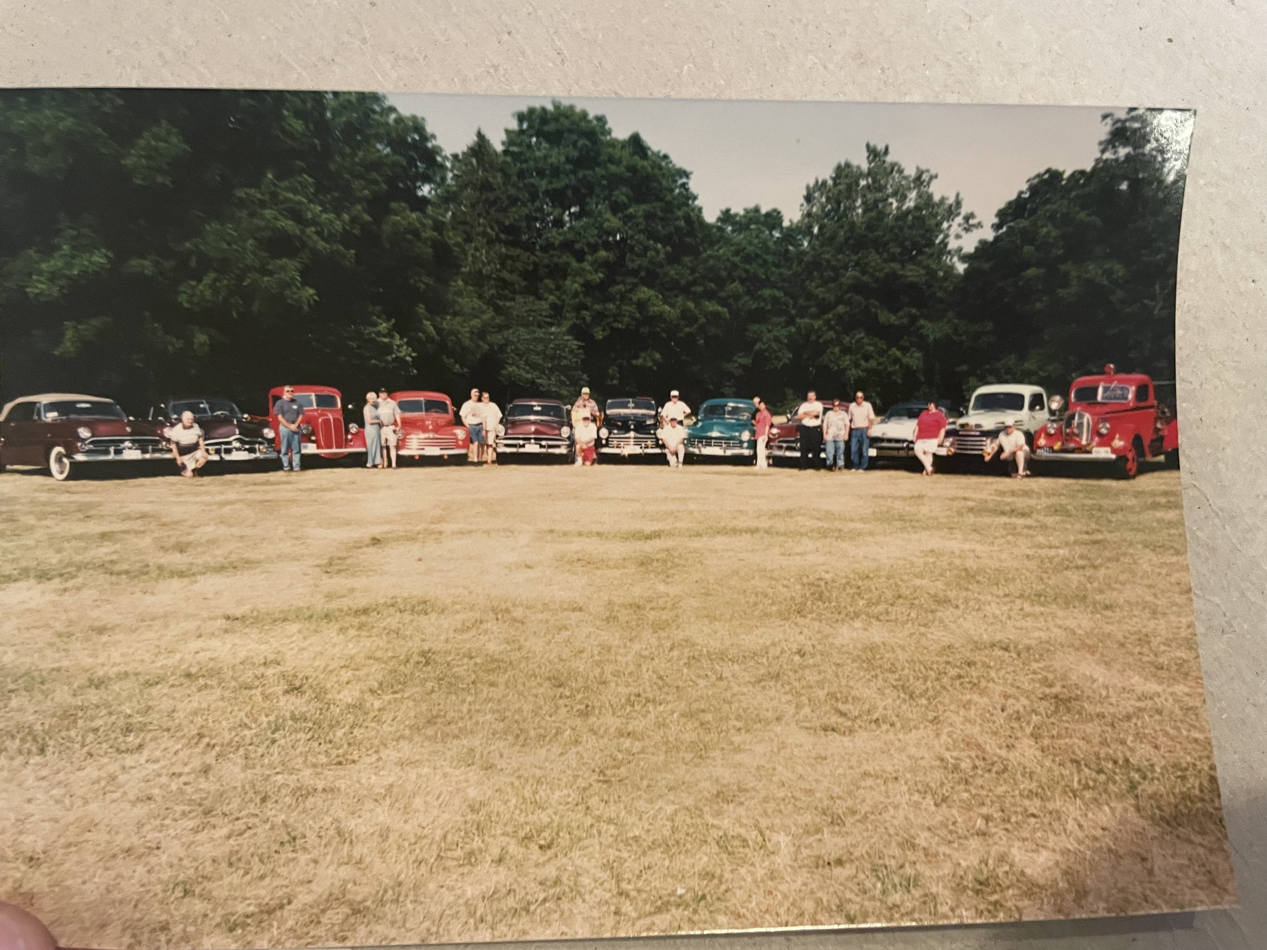 A group of people standing and sitting in front of seven vintage cars parked on grass with trees in the background.