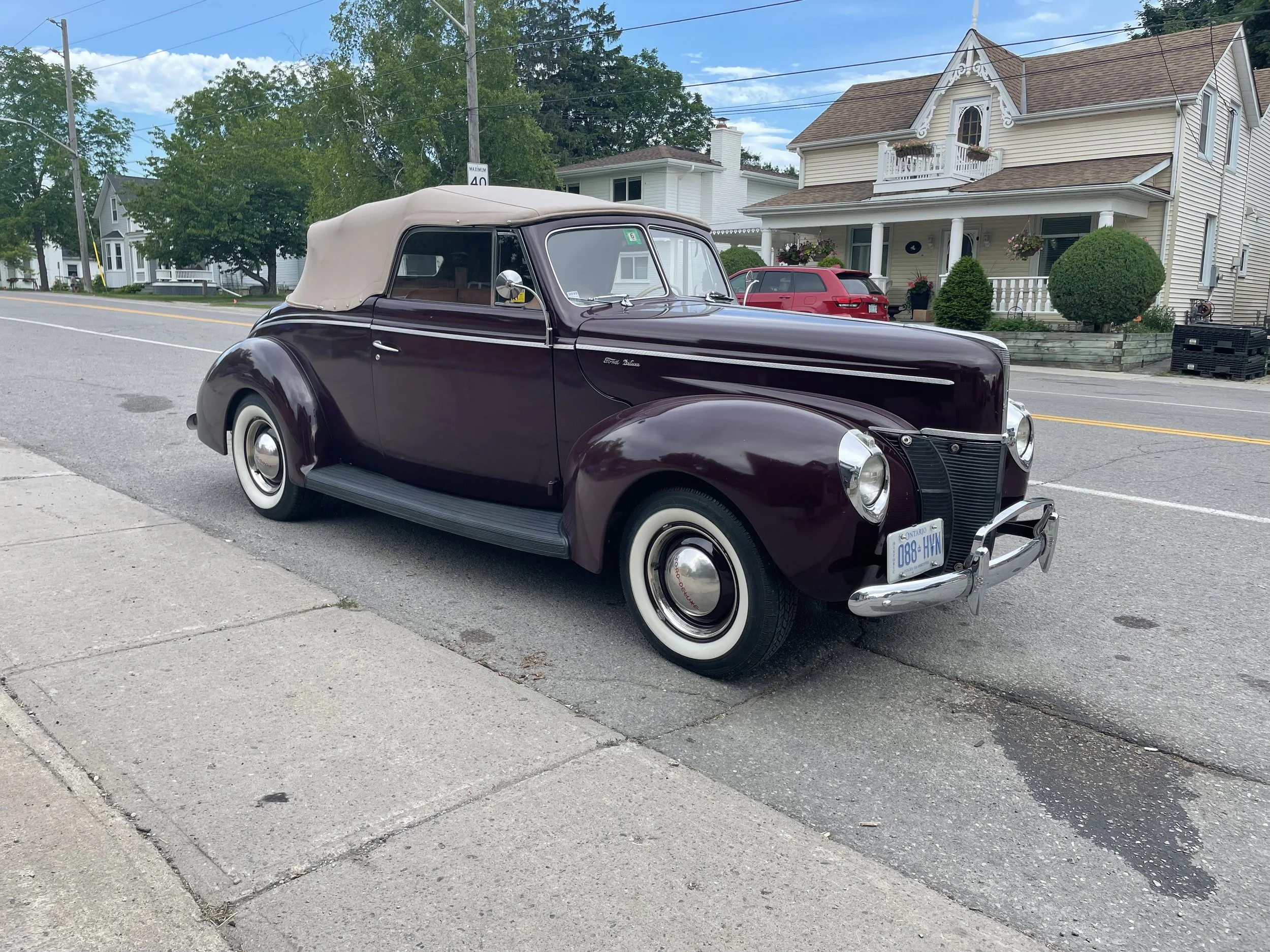 Vintage dark purple convertible car with a beige soft top parked on a city street, with houses and trees in the background.