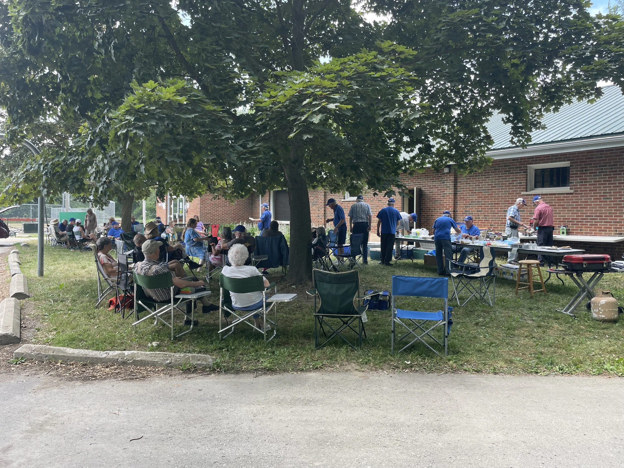 Outdoor gathering with people sitting on chairs under trees and others preparing or serving food near a brick building.