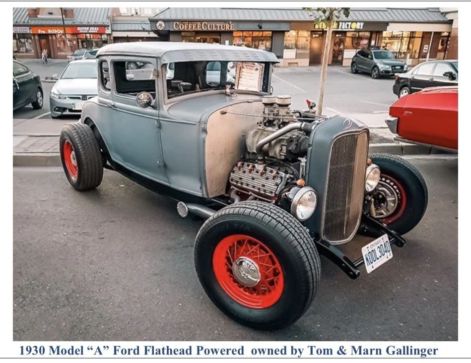 A vintage 1930 Model A Ford flathead hot rod with an unfinished metal hood, red wheels, and an exposed engine, parked on a city street with storefronts and cars in the background.