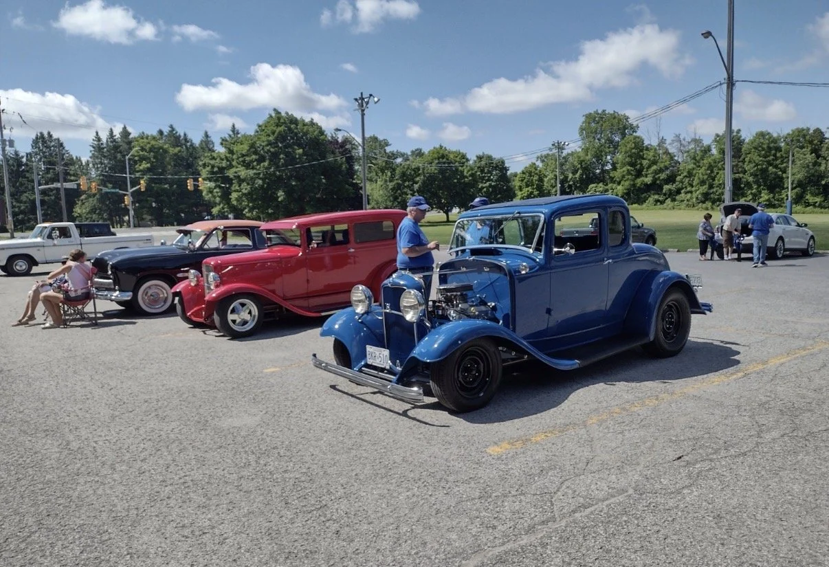 A group of vintage cars parked in a lot on a sunny day, with people inspecting and sitting near them, green trees and a blue sky in the background.