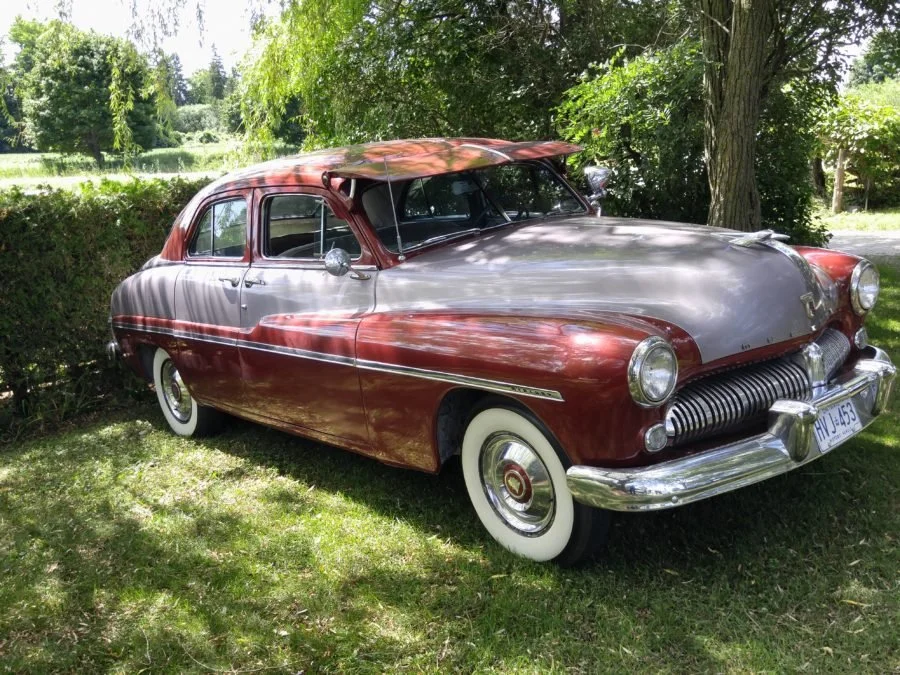 Vintage two-tone red and silver classic car parked outdoors on grass, with trees and bushes in the background.