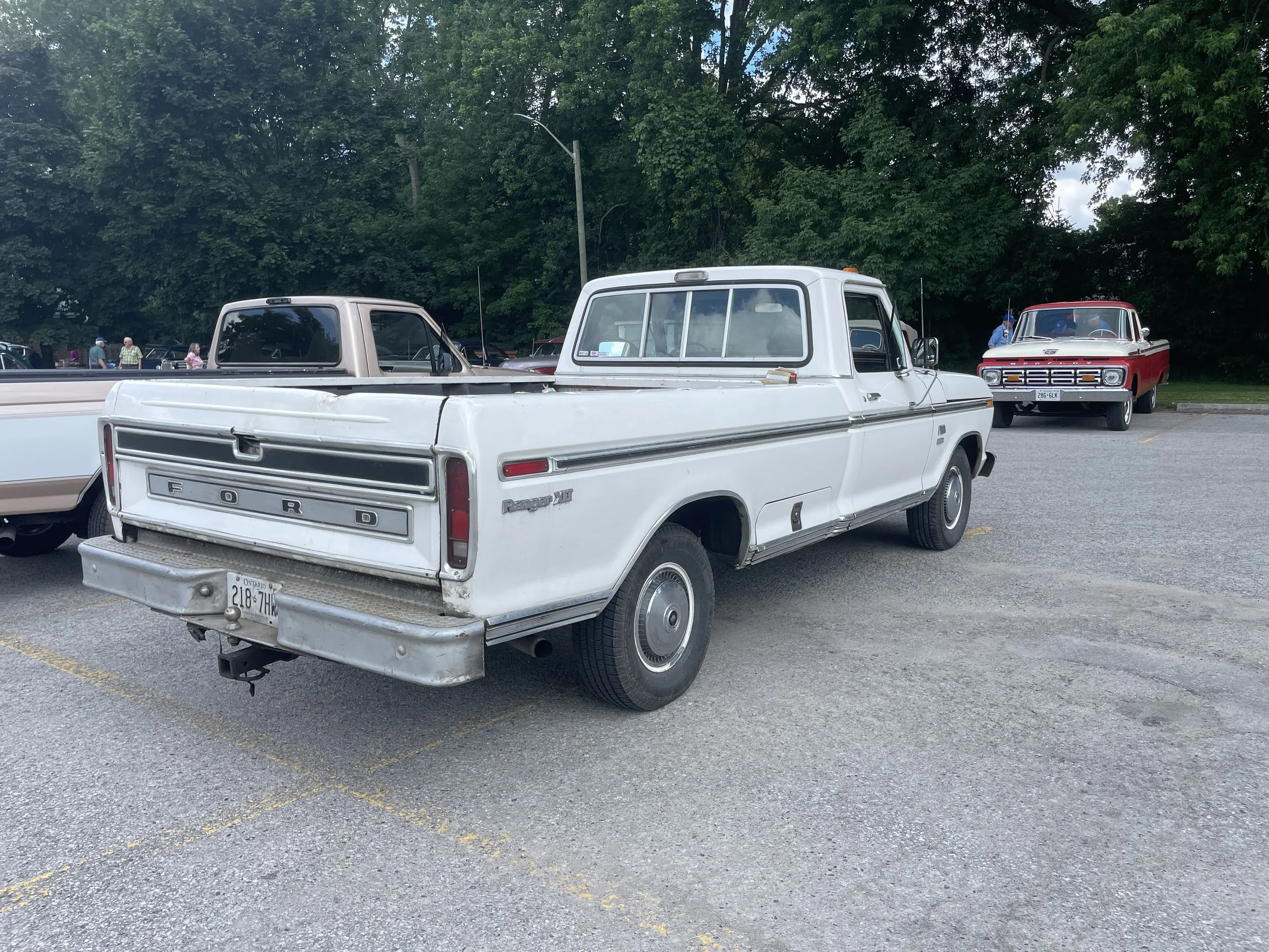 A vintage white Ford Ranger XLT pickup truck parked in a lot, with a red classic car in the background and people walking near trees.