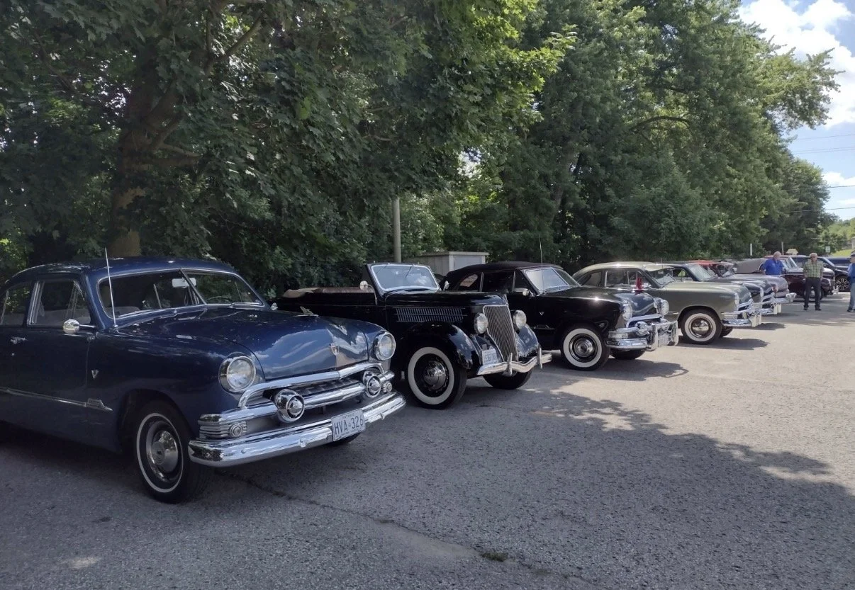 A row of vintage cars parked on a street under trees, with people walking in the background.