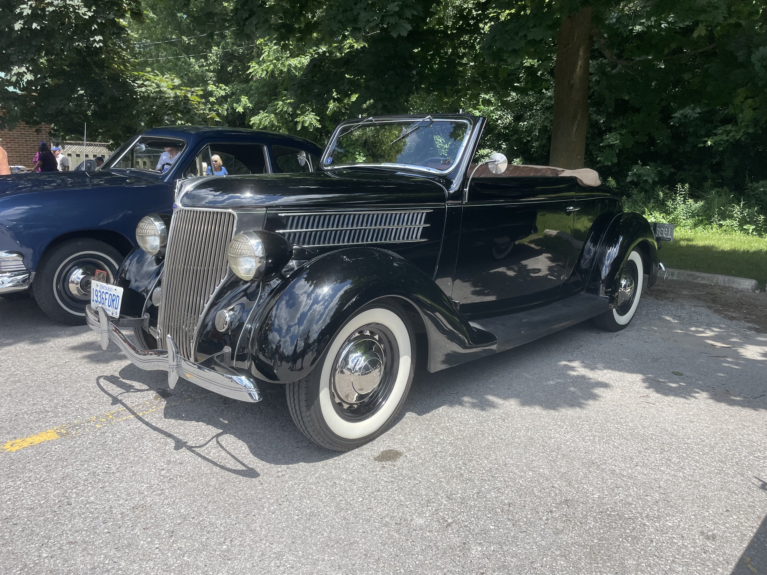 A vintage black convertible car with whitewall tires parked on a gray asphalt surface, with trees and people in the background.