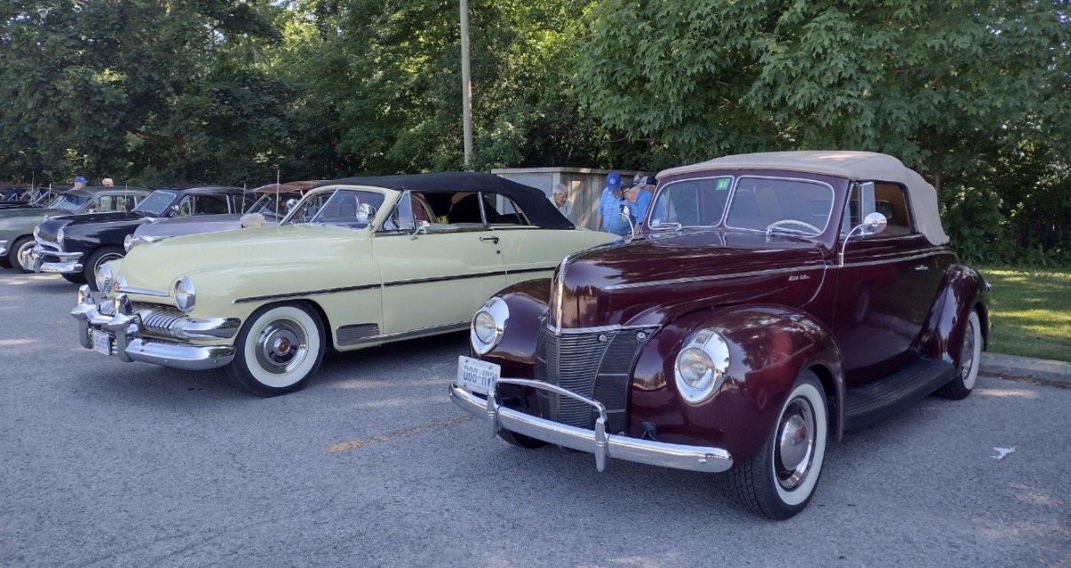 A row of vintage cars parked outdoors with trees in the background, including a cream-colored convertible with black roof and a dark red convertible with tan soft top.