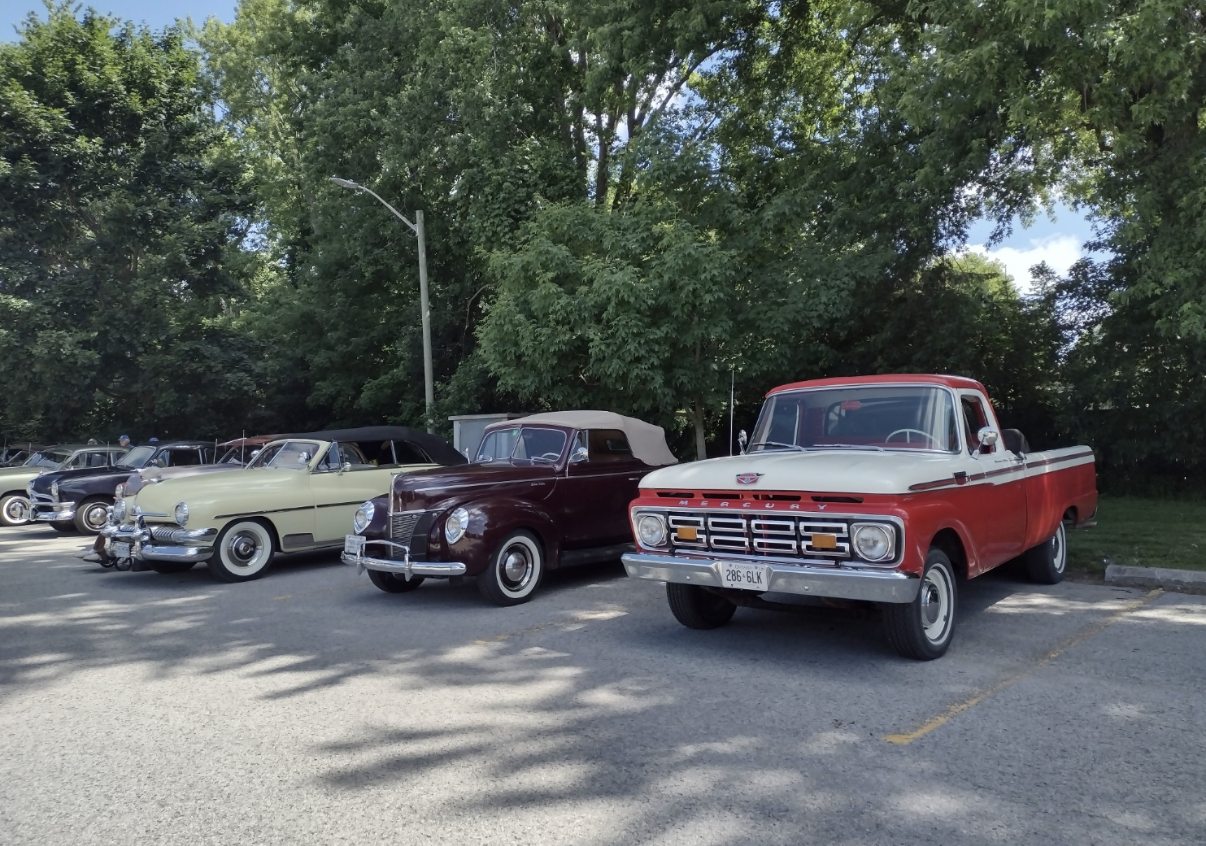 A lineup of vintage cars parked in a lot under trees, including a cream-colored convertible, a burgundy convertible with a beige top, and a red and white pickup truck.