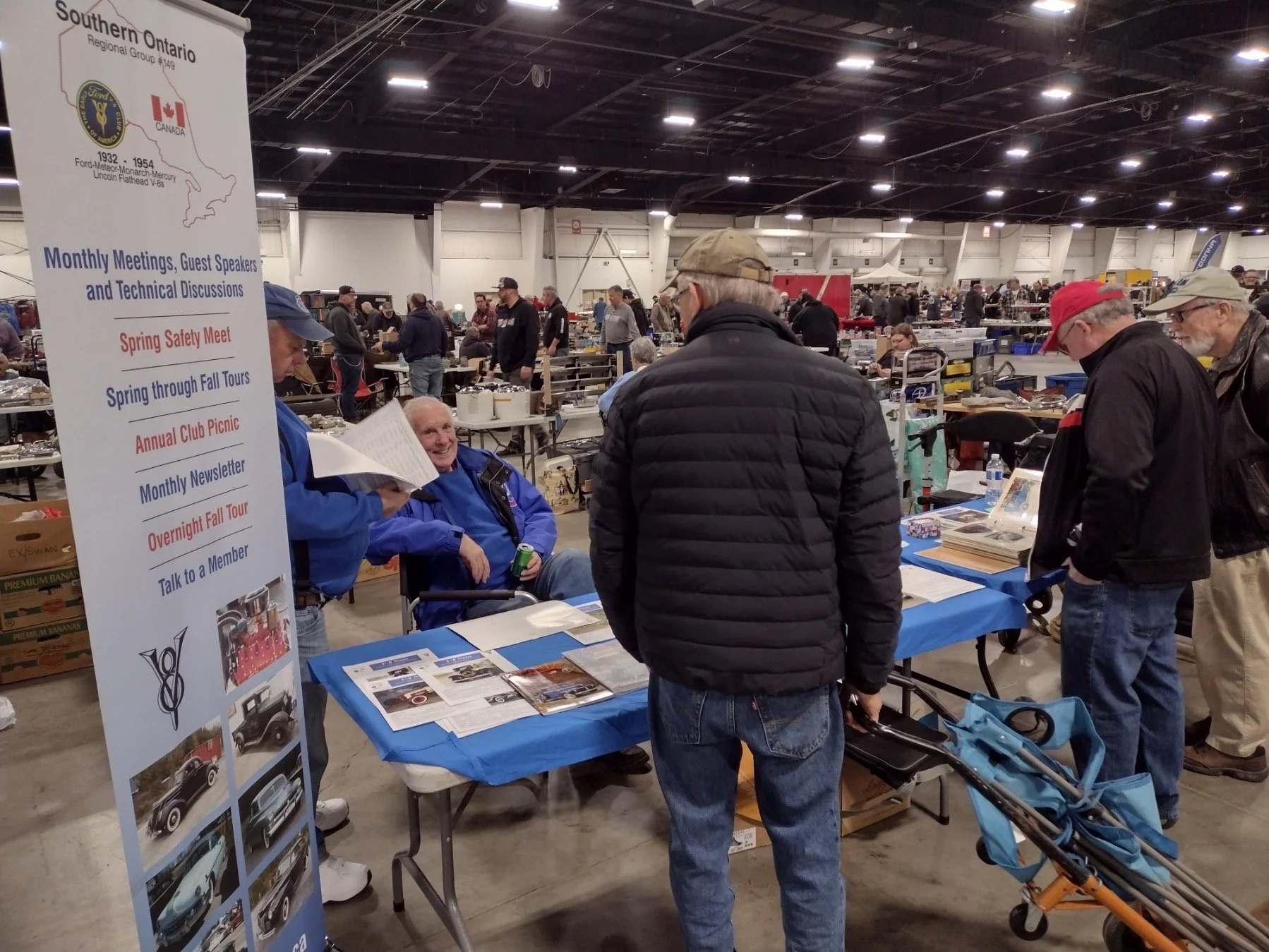People browsing at a vintage car memorabilia sale or expo, with a sign highlighting events for a Southern Ontario region, including monthly meetings and tours, and an on-site table with informational materials.