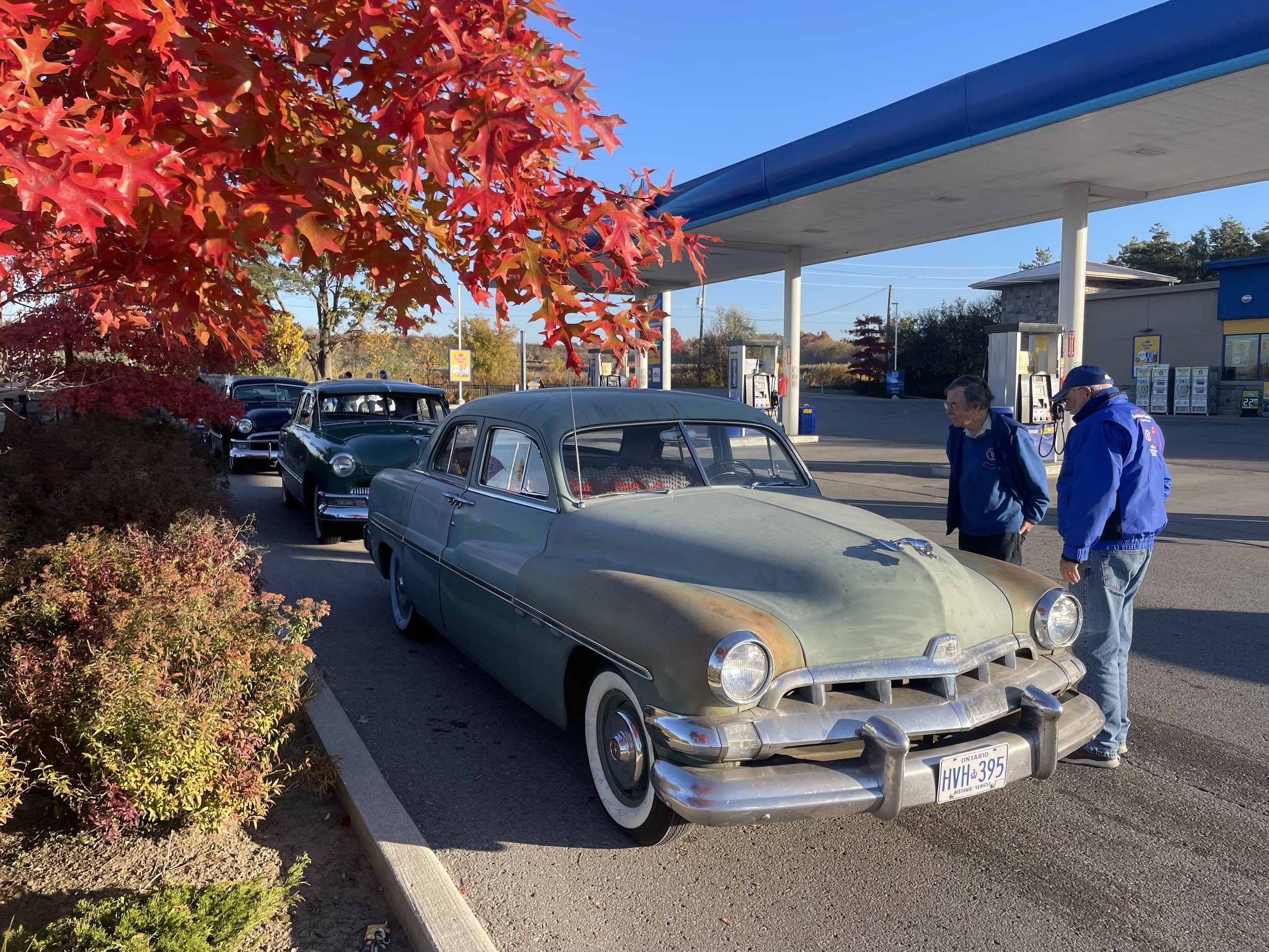 Two men in blue jackets talking near vintage cars at a gas station during fall with colorful red and yellow leaves on trees.