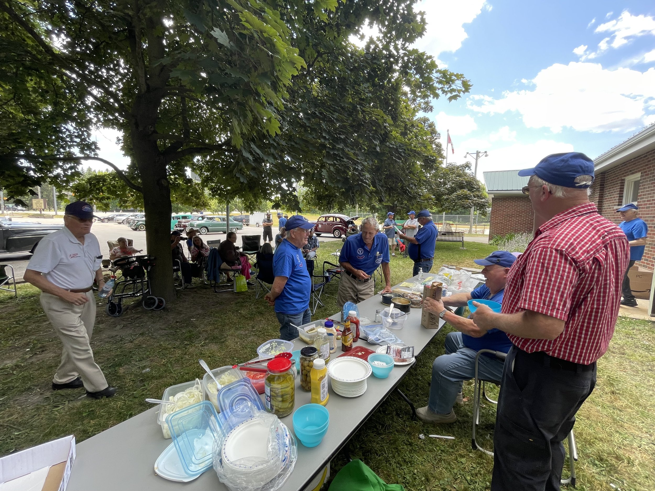 A group of people gathers outdoors under a large tree on a sunny day, with some sitting on chairs and others standing around tables with food and condiments, near a brick building and a parking lot.