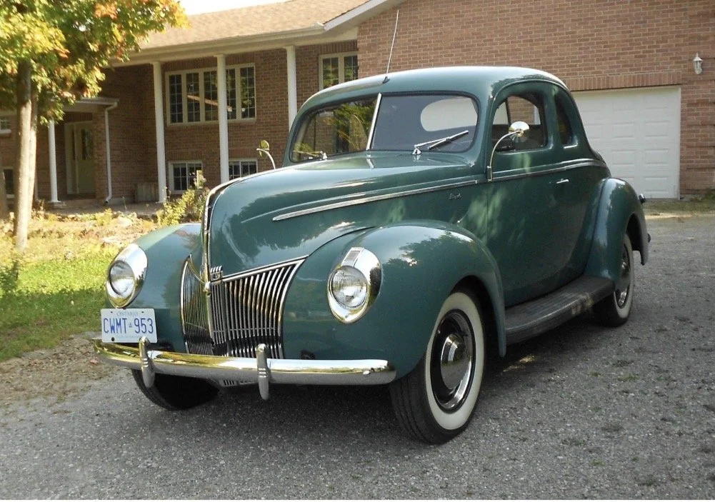 A vintage green car parked on a driveway in front of a brick house with white trim and a garage door.
