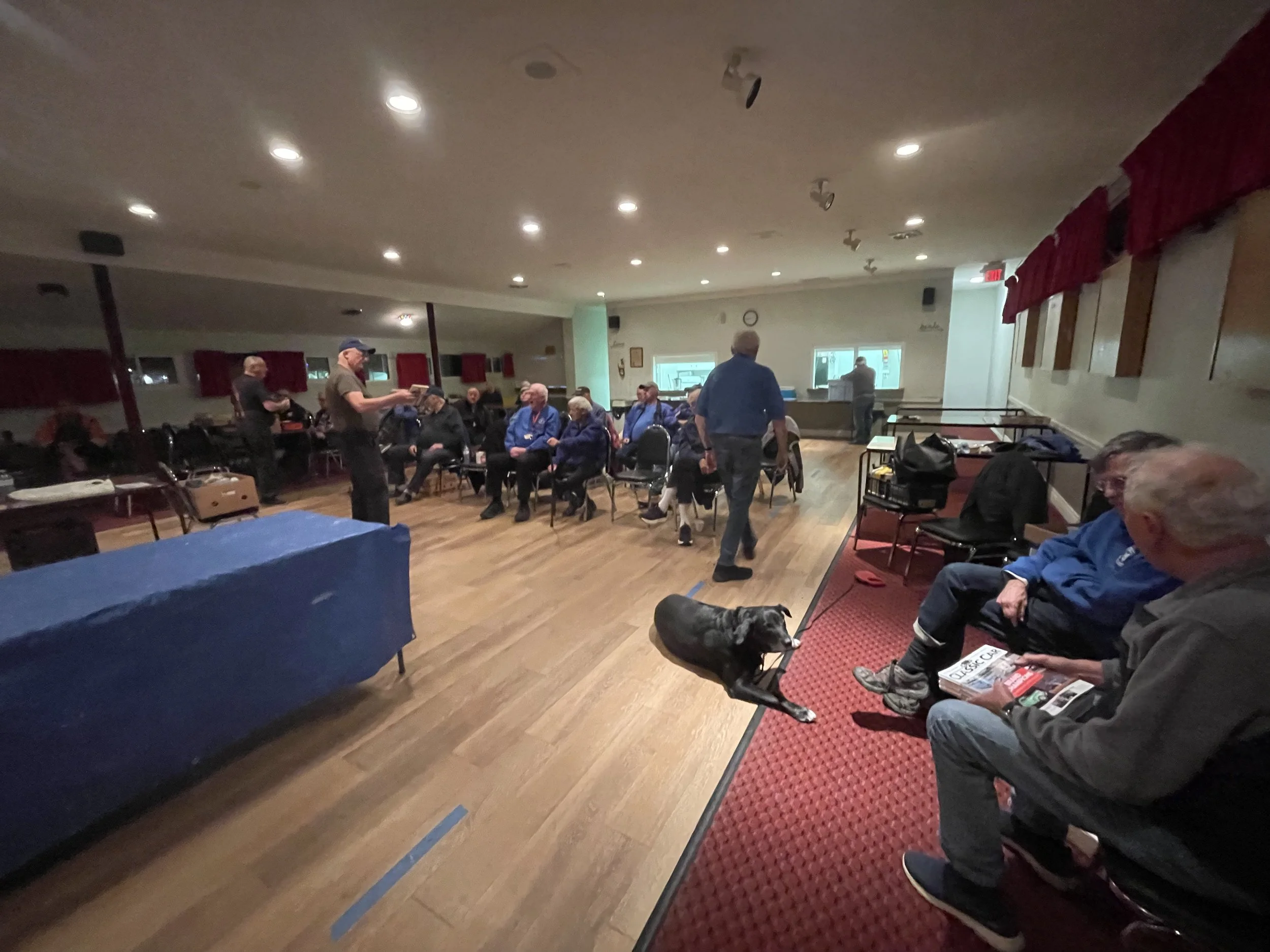 People gathered in a room with wooden floors, some sitting in chairs and others standing, while two men appear to be preparing or reading something; a dog is lying on the red carpeted floor near two seated men, one of whom is holding magazines, with 