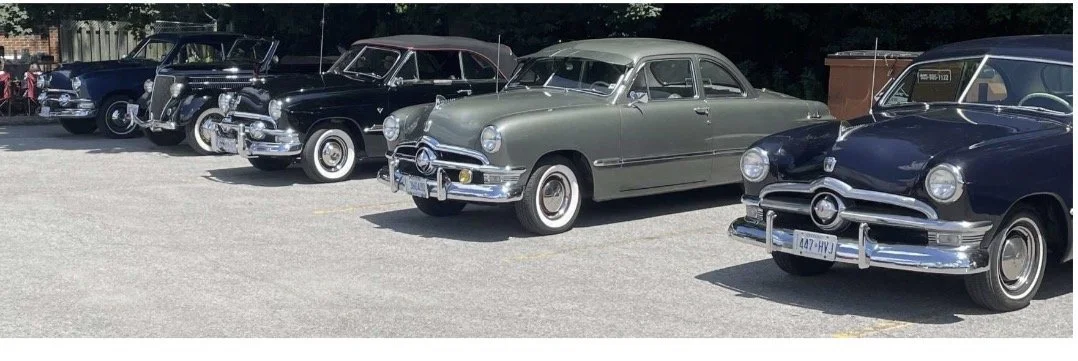 A row of vintage cars parked in a lot, including black, gray, and blue models from the mid-20th century.