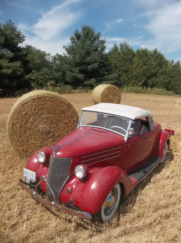 A vintage red convertible car with a white roof parked on a hayfield, with large round hay bales and trees in the background.
