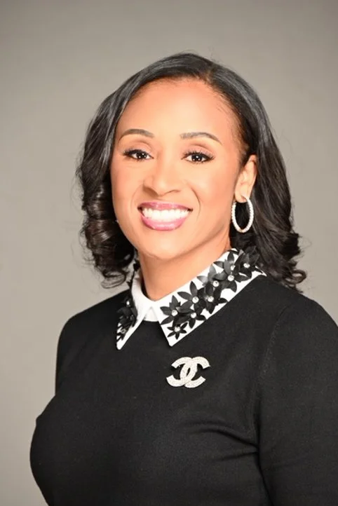 Professional woman with black, curly hair, wearing a black top with a white and black patterned collar, silver hoop earrings, and a Chanel logo pin, smiling at the camera.