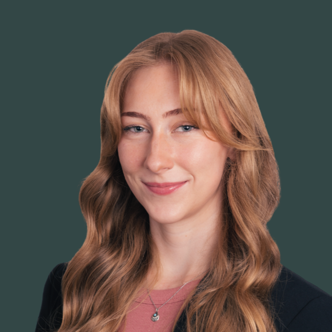 A woman with long wavy ginger hair, wearing a black blazer and a pink top, smiling at the camera against a dark teal background.