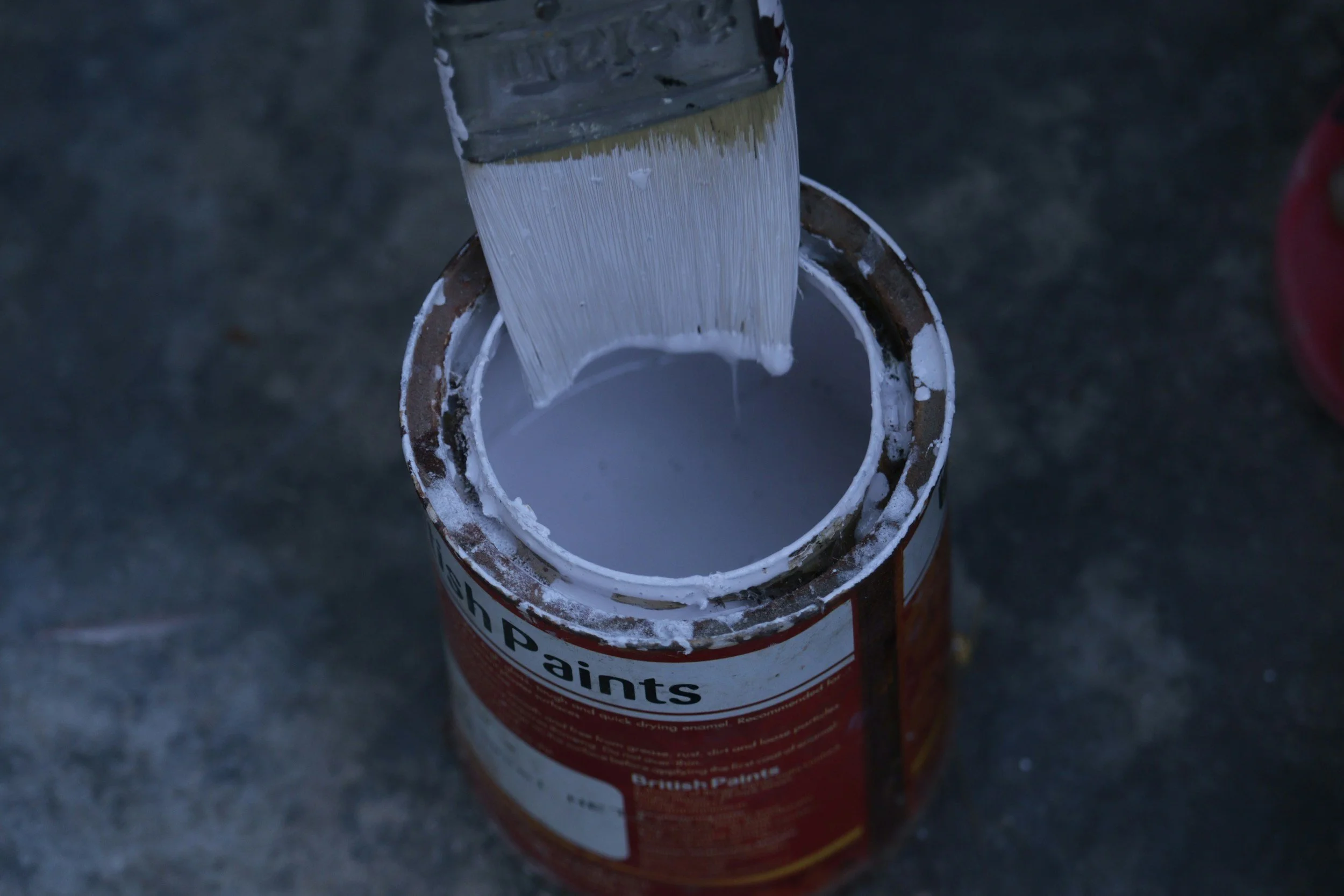 A paintbrush dipping into a can of white paint, with some paint on the rim, on a dark surface.