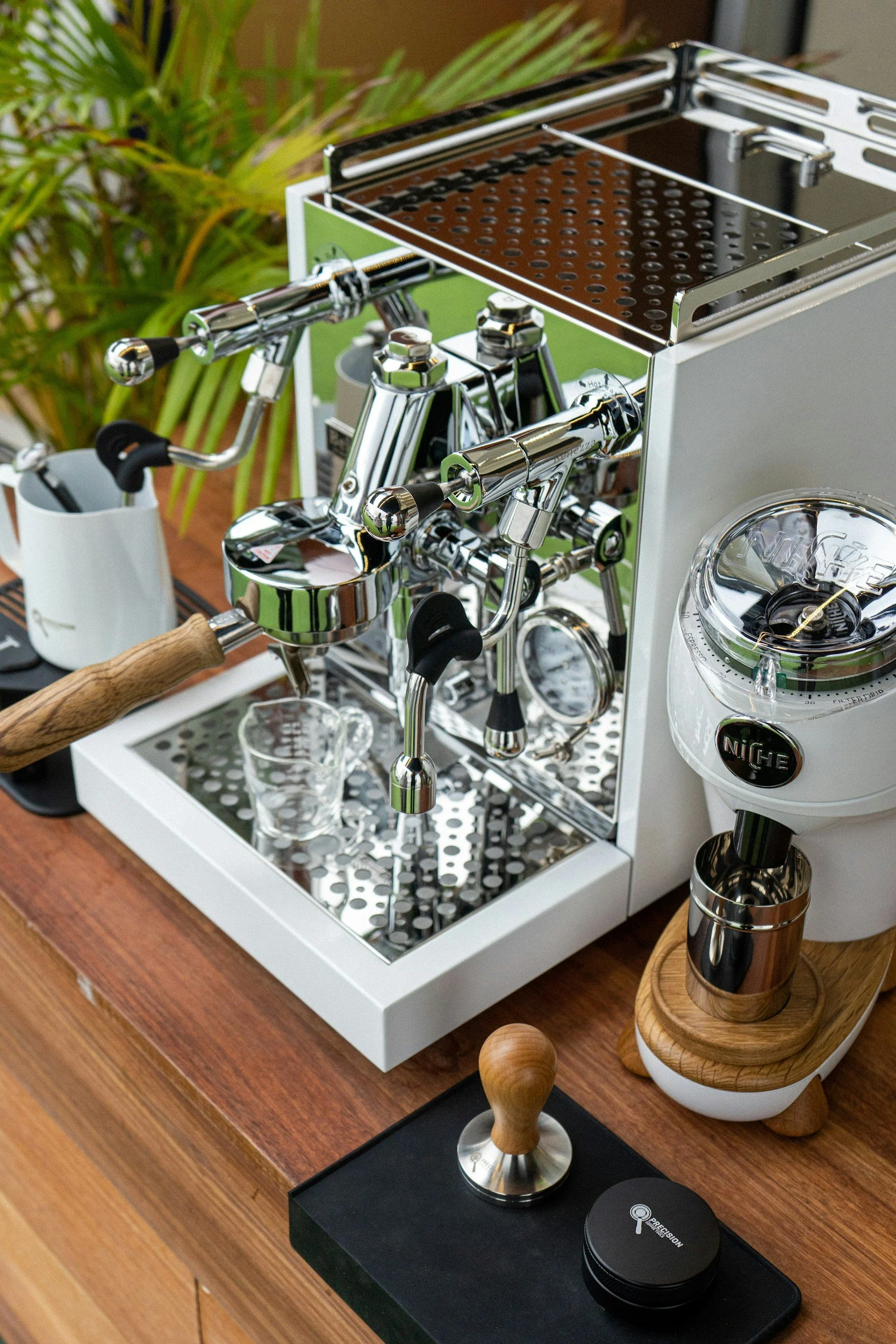 A close-up of a professional espresso machine with chrome and black fittings on a wooden counter, with various coffee accessories including a tamper, coffee grinder, and a coffee scoop on a black stand, surrounded by green plants in the background.