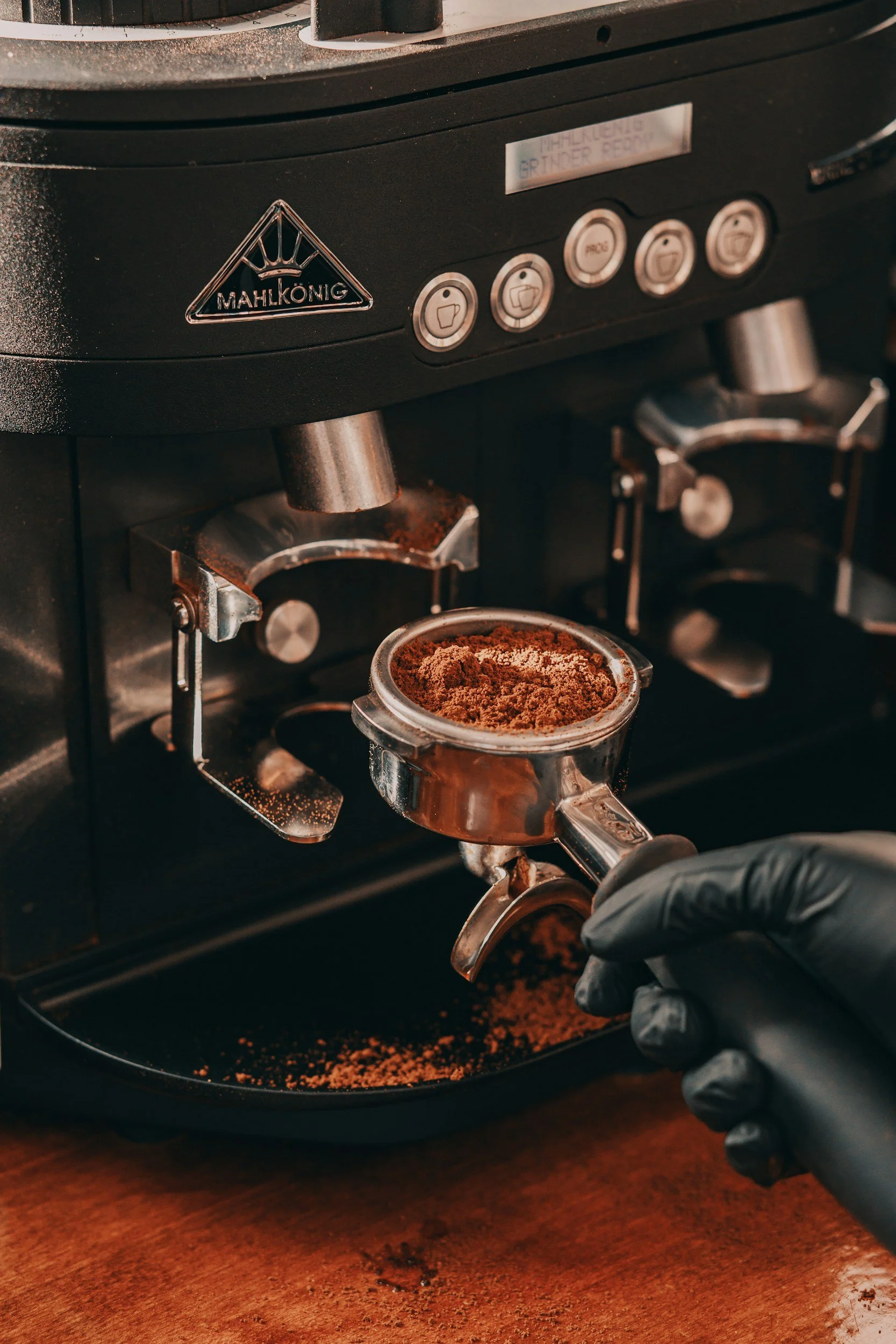 Close-up of a barista pressing ground coffee into a portafilter underneath a Mahlkönig coffee grinder, with spilled coffee grounds on a wooden countertop.