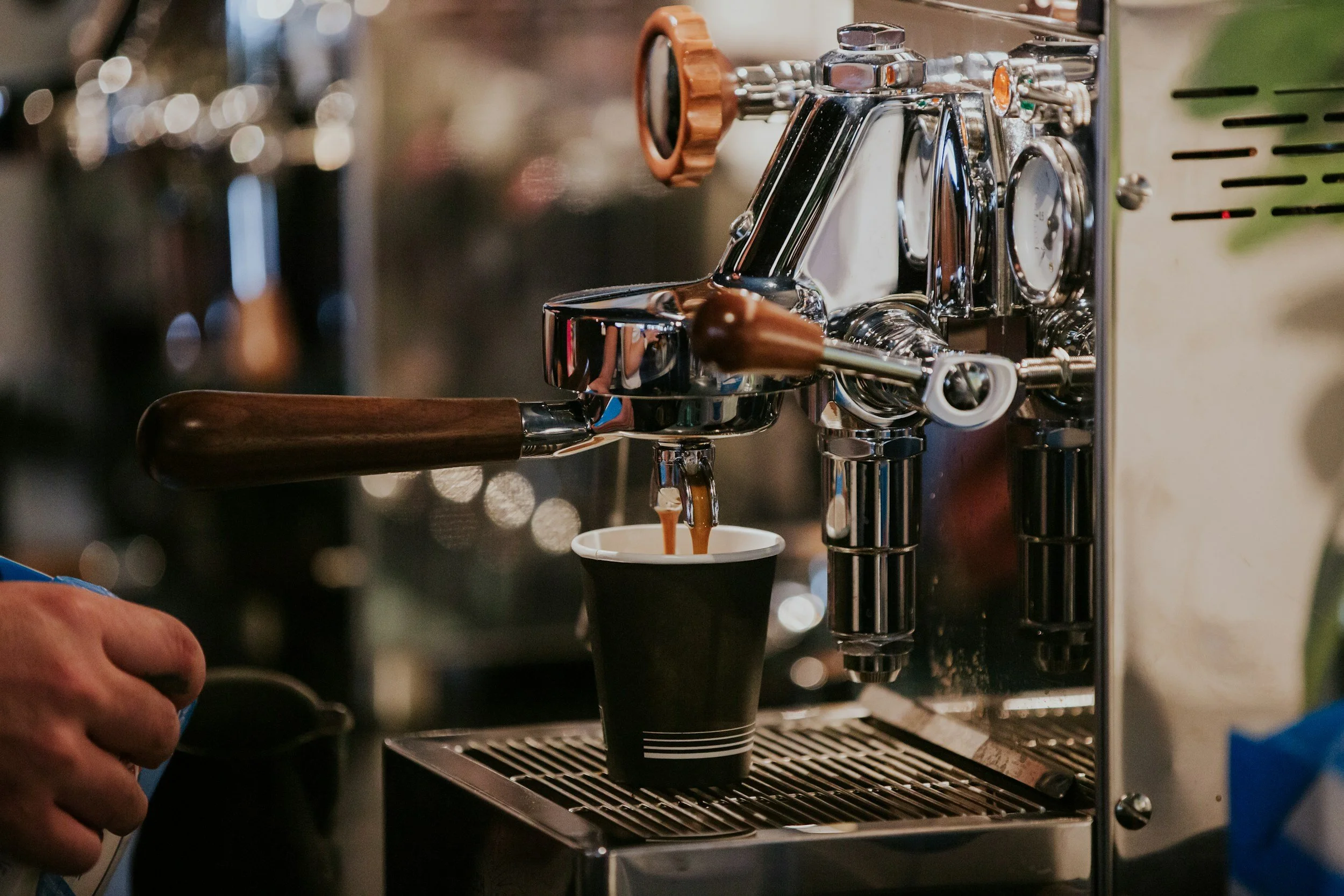 A close-up of a coffee espresso machine with a portafilter attached, pouring coffee into a small paper cup.
