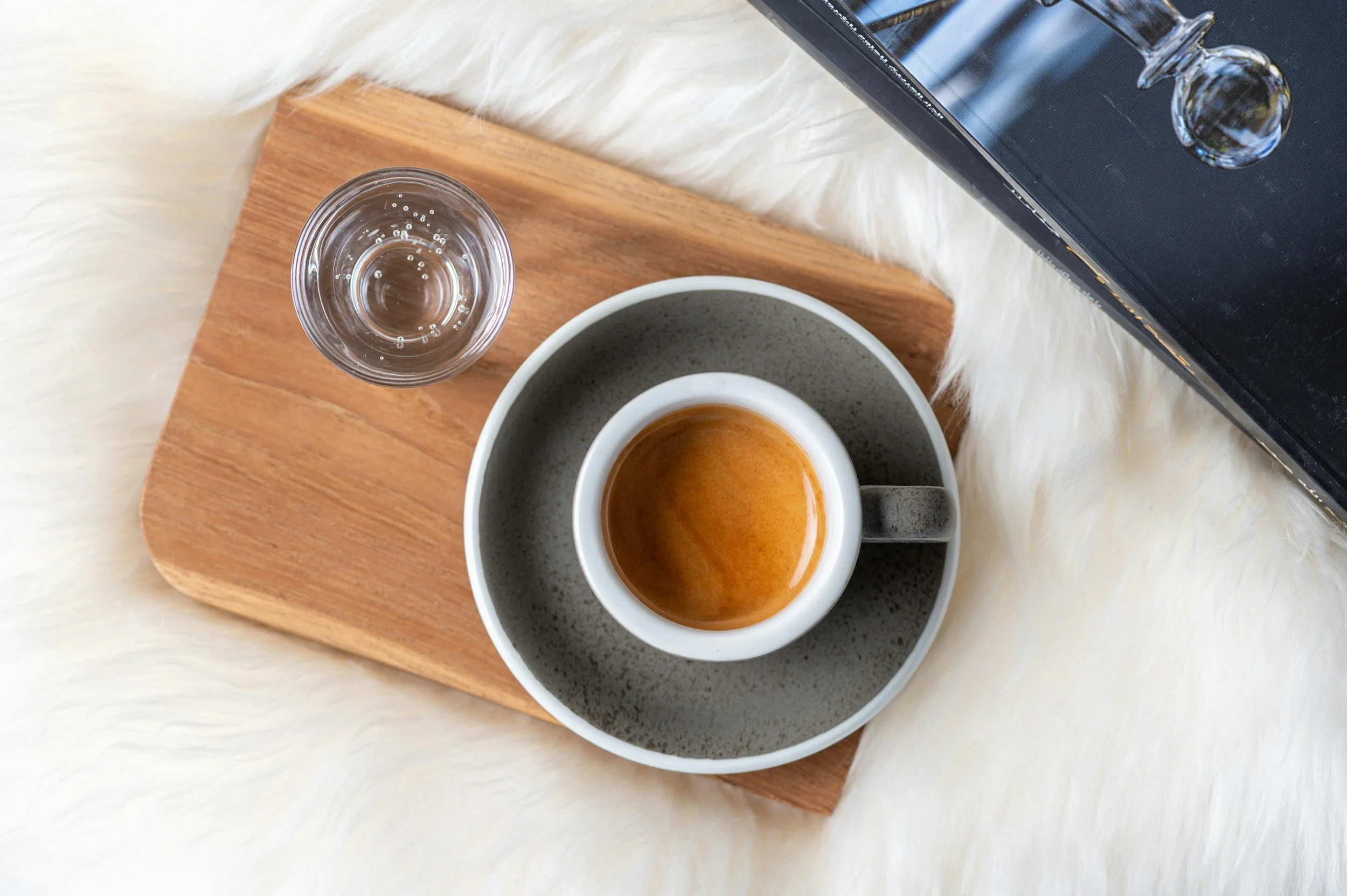A top view of a small cup of espresso, a glass of water, a wooden tray, and a black laptop on a white furry surface.