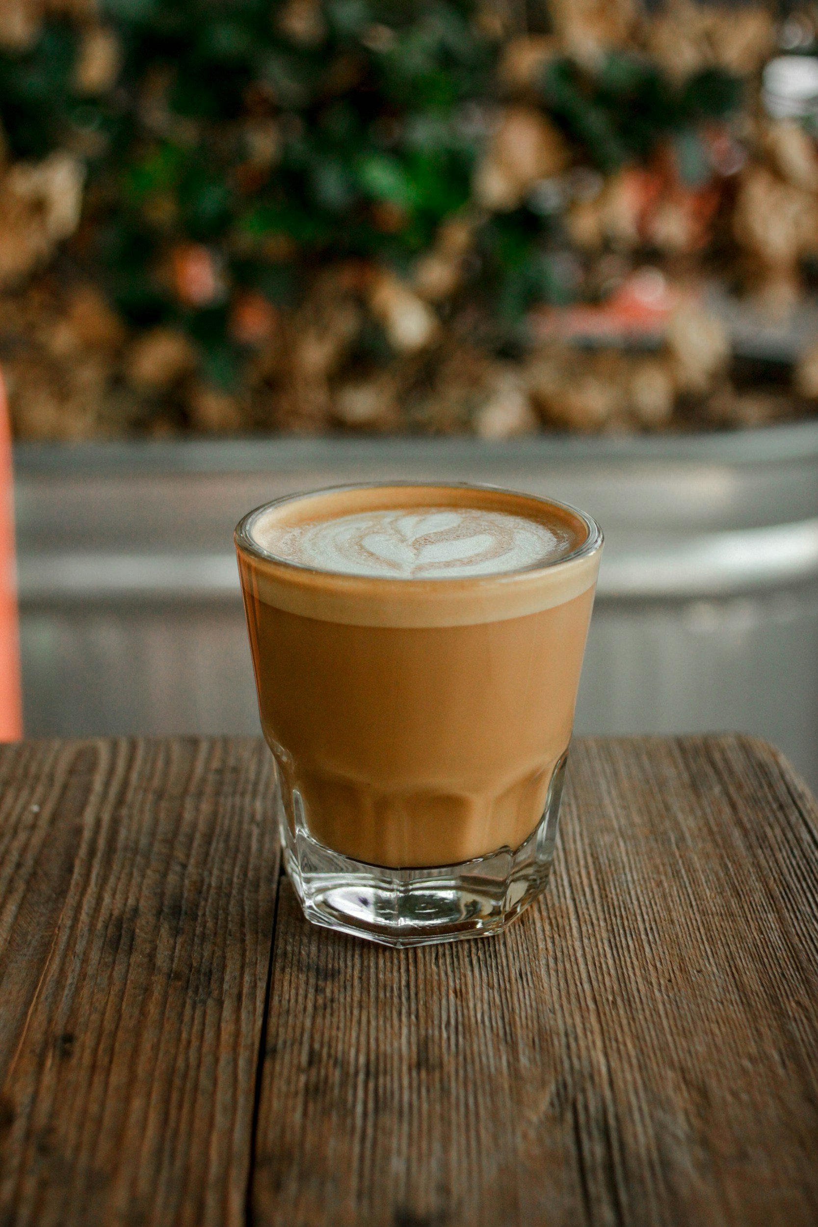 A glass of coffee with a swirl of frothy milk on top, sitting on a wooden surface, with blurred background of plants and rocks.