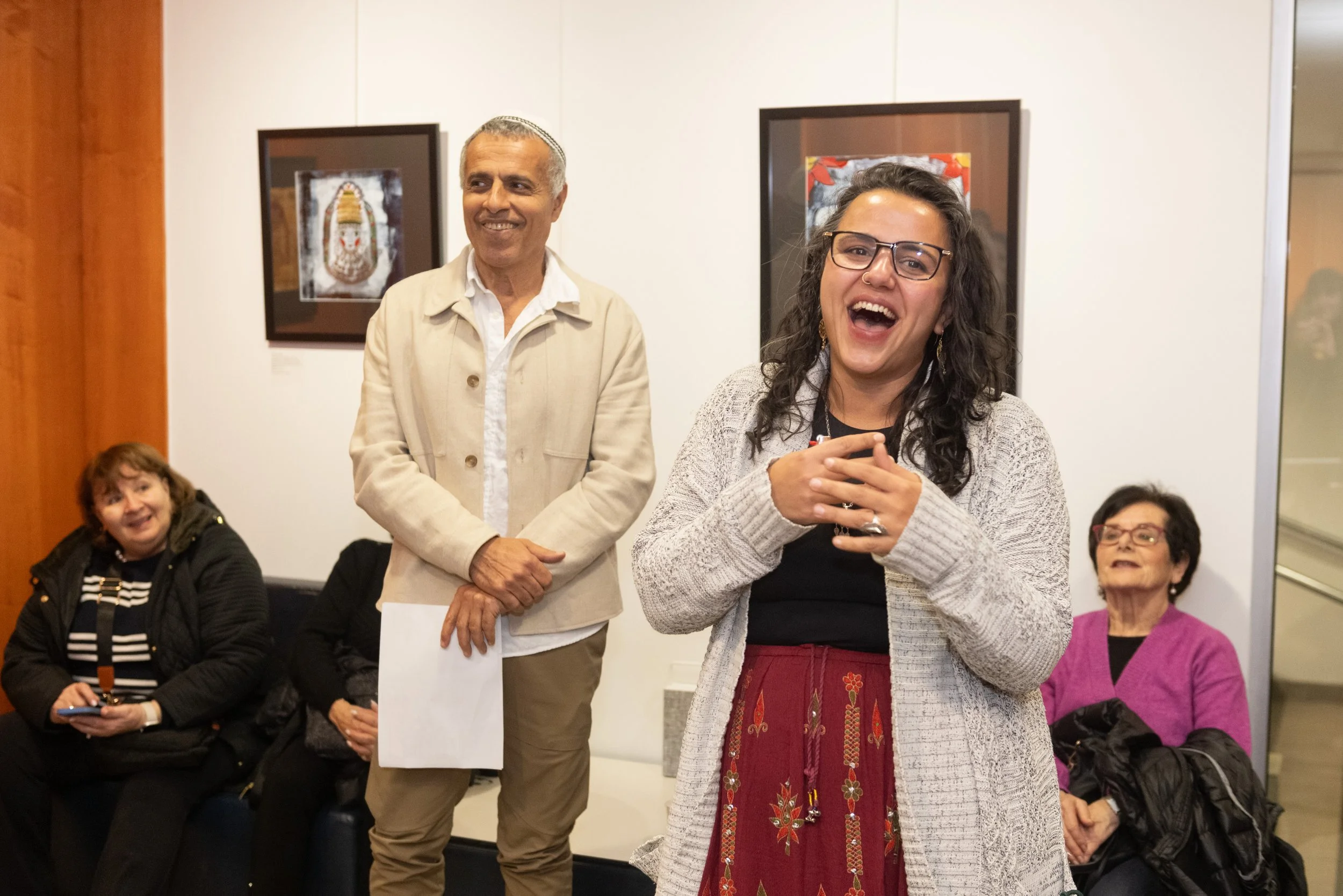 Group of four people at an indoor social gathering, woman in front laughing and holding a cup, three others sitting or standing behind her, framed artwork on white walls