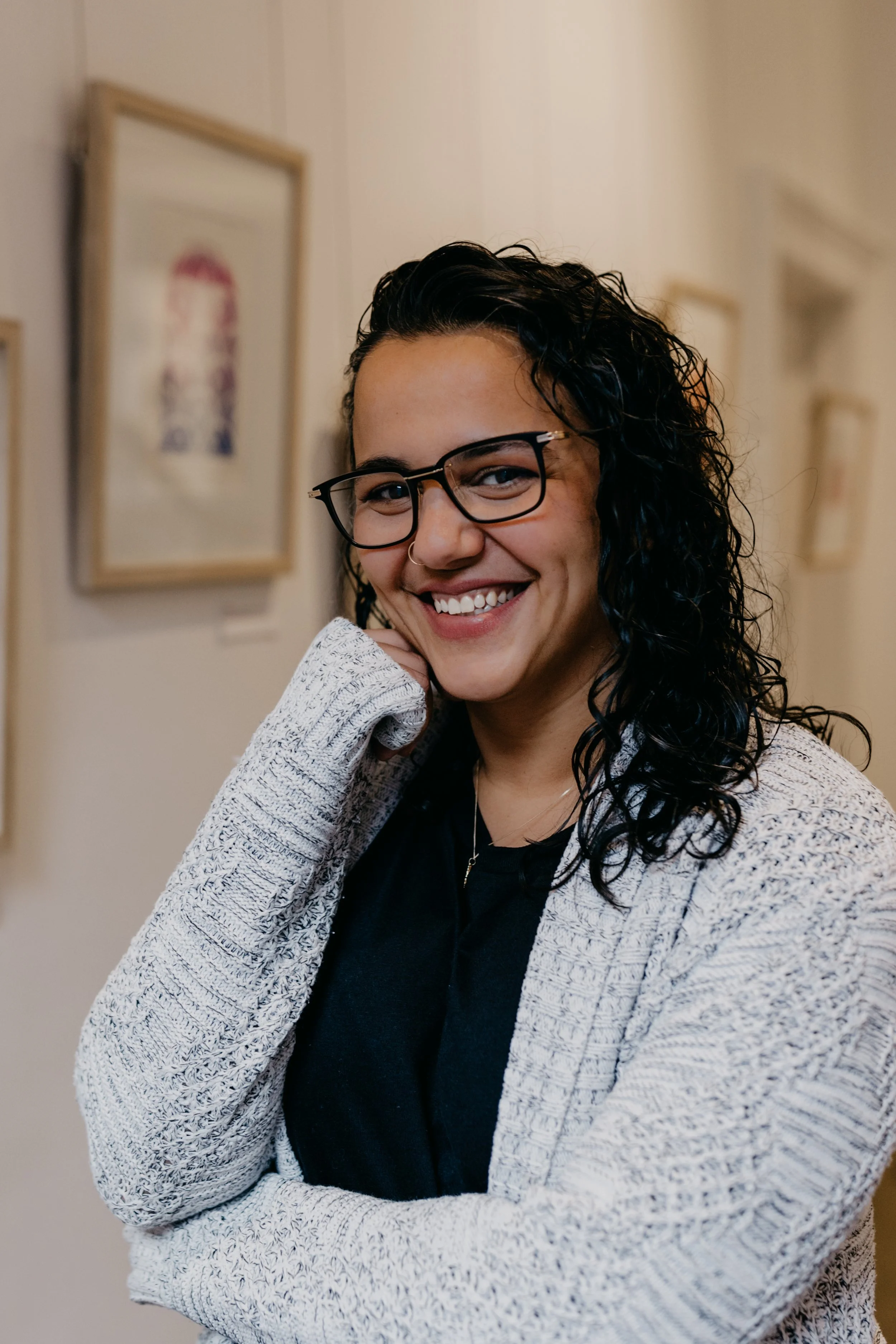 A woman with curly black hair, glasses, and a nose ring, smiling at the camera in an art gallery.