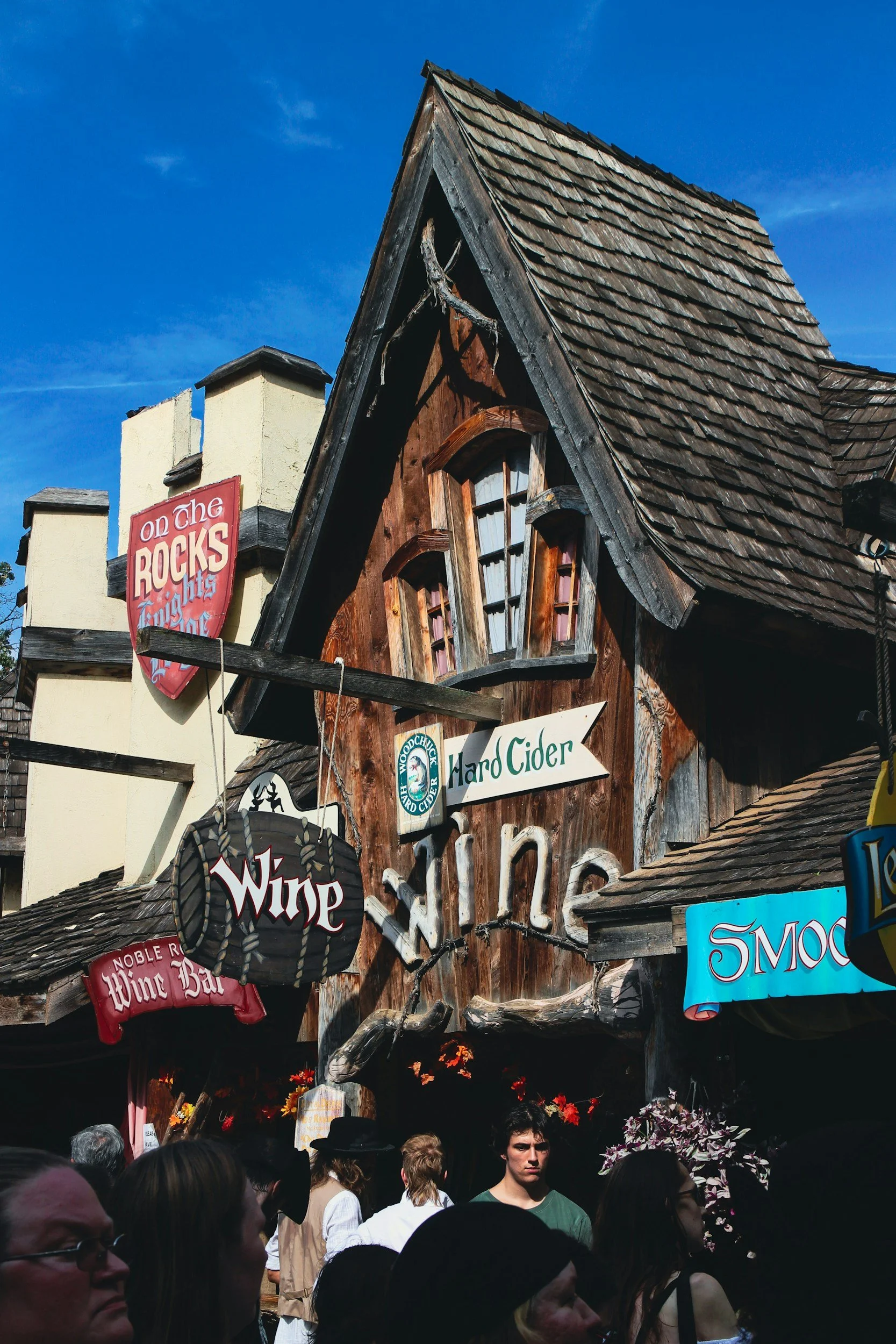 People gathered in front of a rustic wooden building with signs for wine, hard cider, and wine bar, under a sunny sky at a ren faire, renaissance festival.