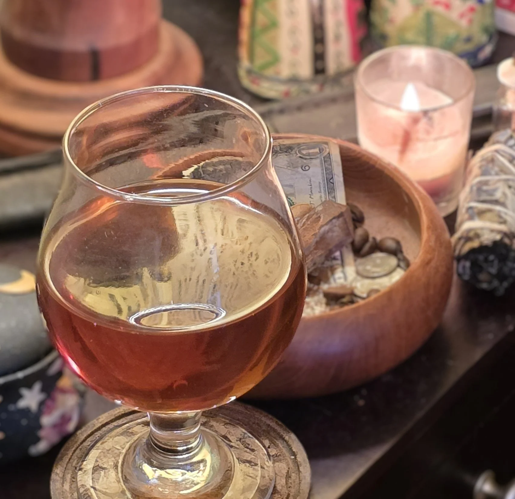 A glass of mead on a wooden coaster with a wooden offering bowl and a $1 bill inside, surrounded by candles and decorative objects.