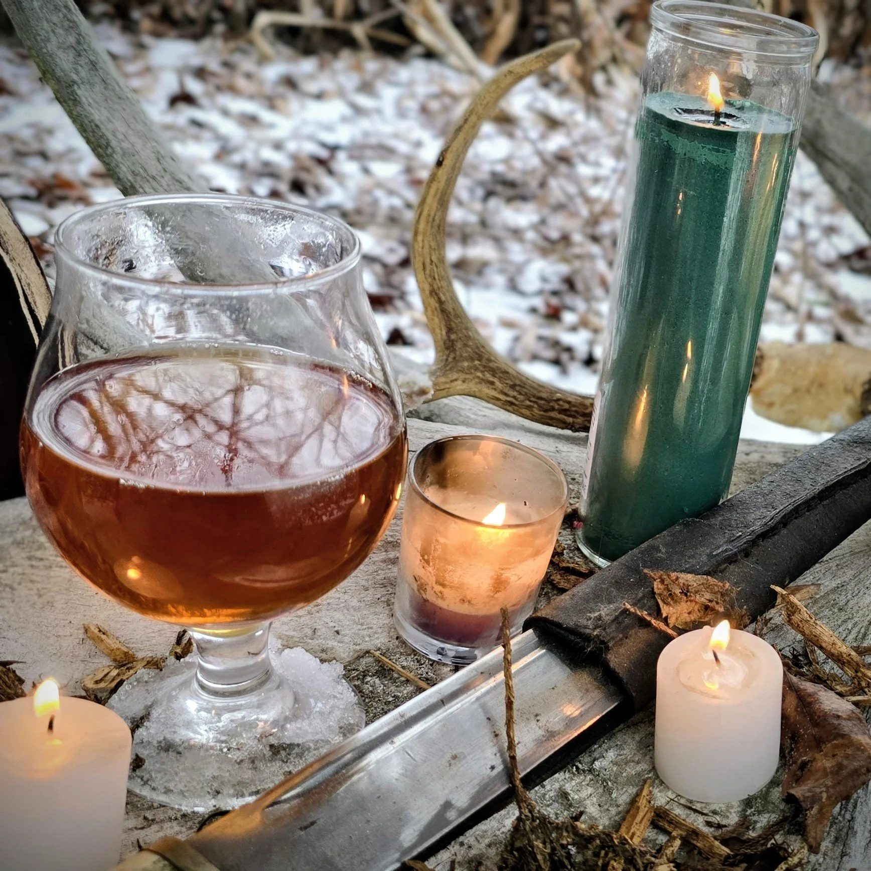 A scene with lit candles, a glass of mead , and a tall green column candle on a rustic surface outdoors, with snow and tree branches in the background, with antlers and altar pieces.