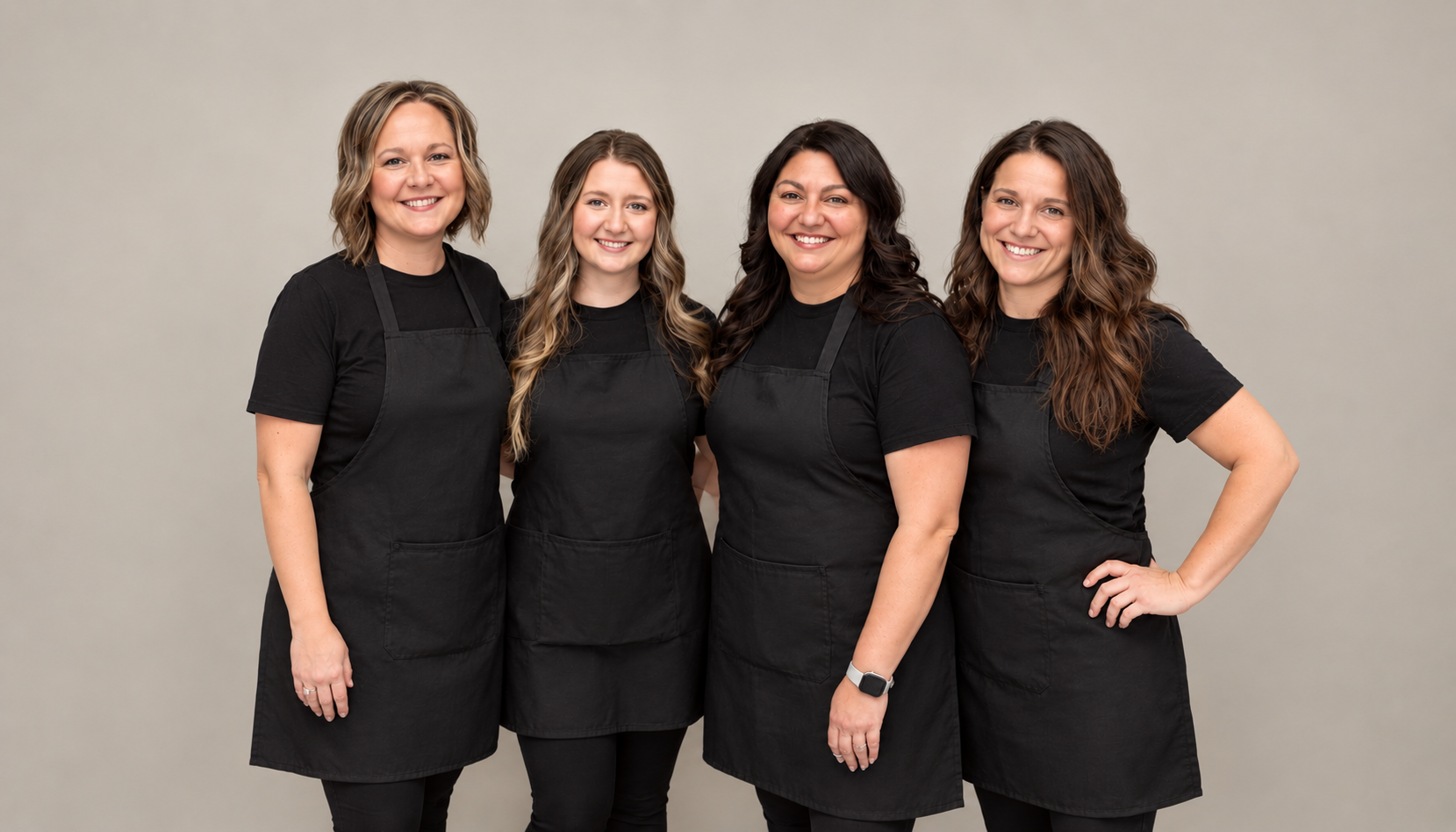 Four women standing together wearing black t-shirts and black aprons, smiling at the camera against a plain background.