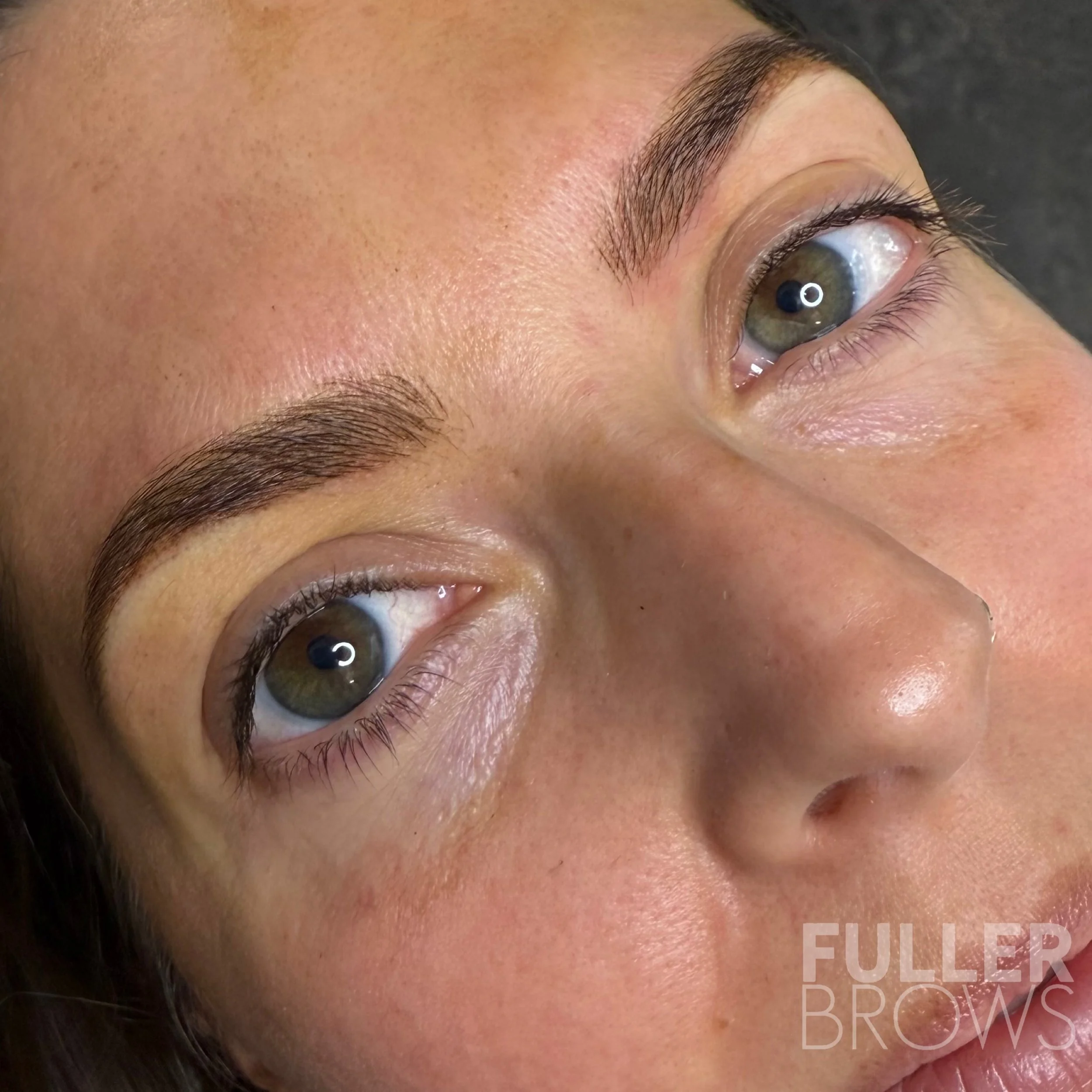 Close-up of a person's face focusing on well-groomed eyebrows and hazel eyes with natural makeup, lying on a dark surface with the text 'FULLER BROWS' in the corner.
