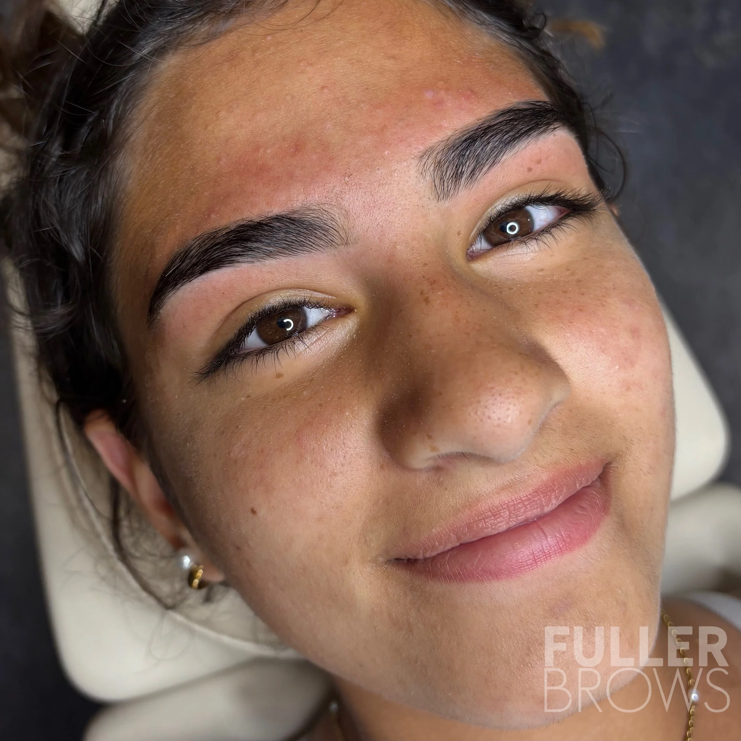 Close-up of a woman with well-defined eyebrows, brown eyes, and a slight smile, lying down with her head tilted slightly. The background is blurred, and there is a watermark that says 'Fuller Brows' in the lower right corner.