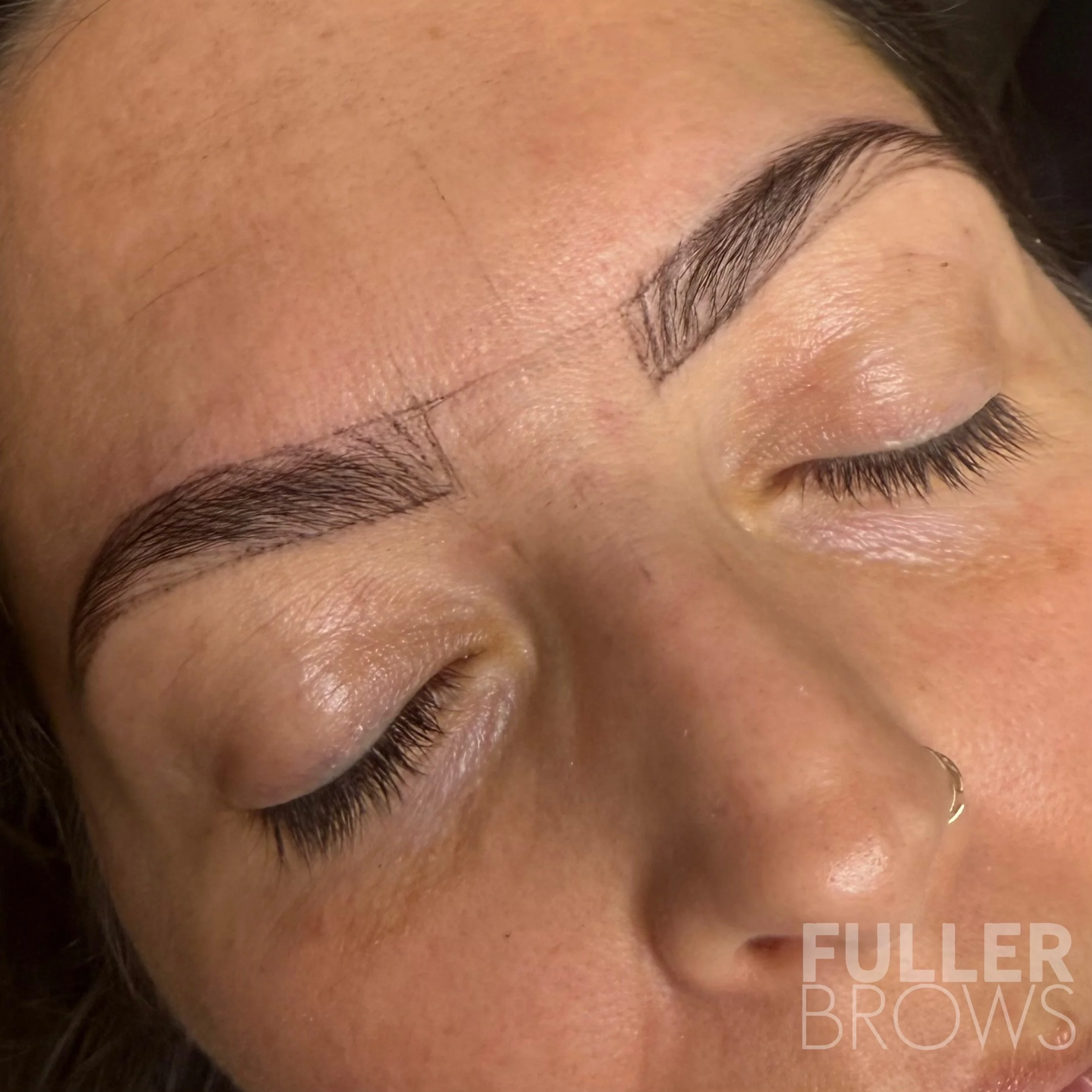 Close-up of a woman’s face with her eyes closed, showcasing freshly shaped and filled eyebrows, and a small nose ring.