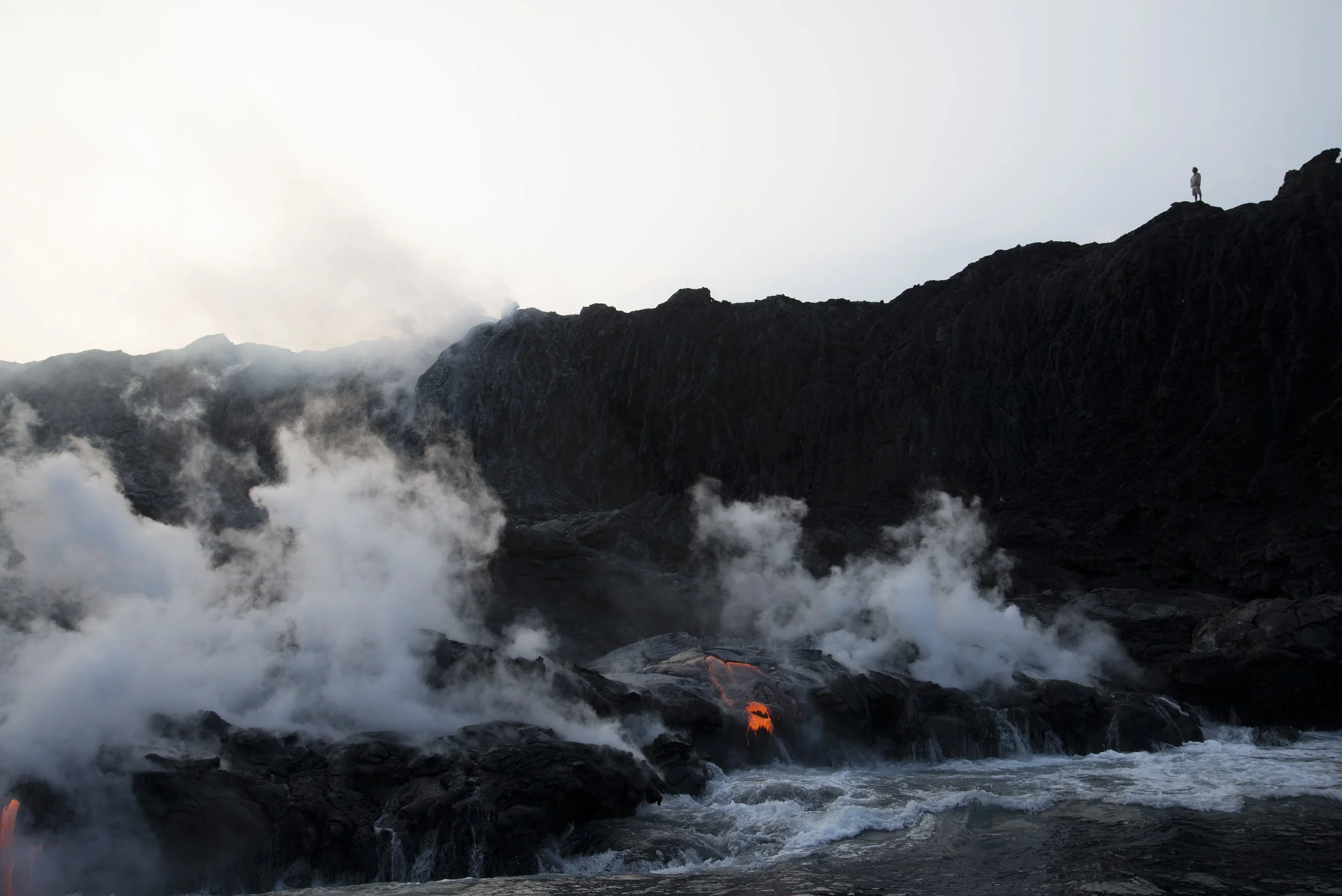 A person standing on dark volcanic rocks above a river of flowing lava with steam and smoke rising around it, in a volcanic landscape.