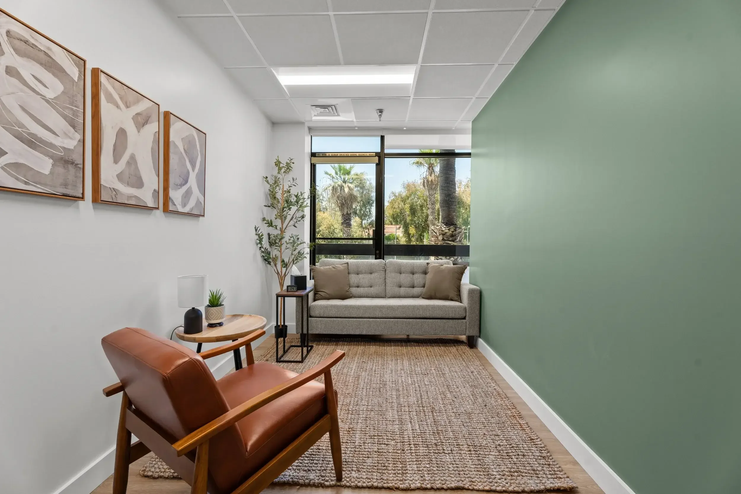 A photo of Dr. Charles' office in Santa Monica. This is a modern room with a grey sofa, a leather armchair, a small side table with a lamp and plant, green accent wall, framed artwork, and large window showing palm trees outside.
