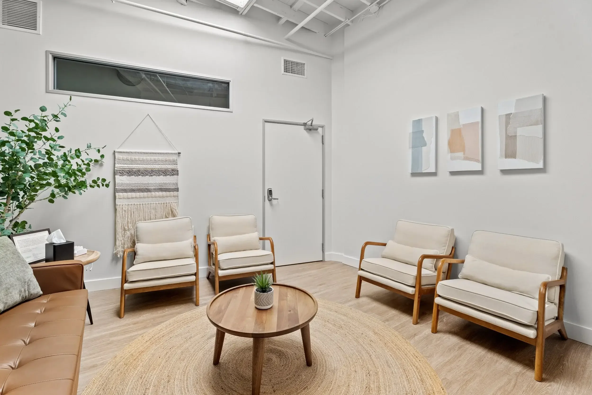 A photo of Dr. Charles' waiting room in Santa Monica. The room has beige armchairs and a tan leather sofa, white walls decorated with abstract art, and a tall green plant.