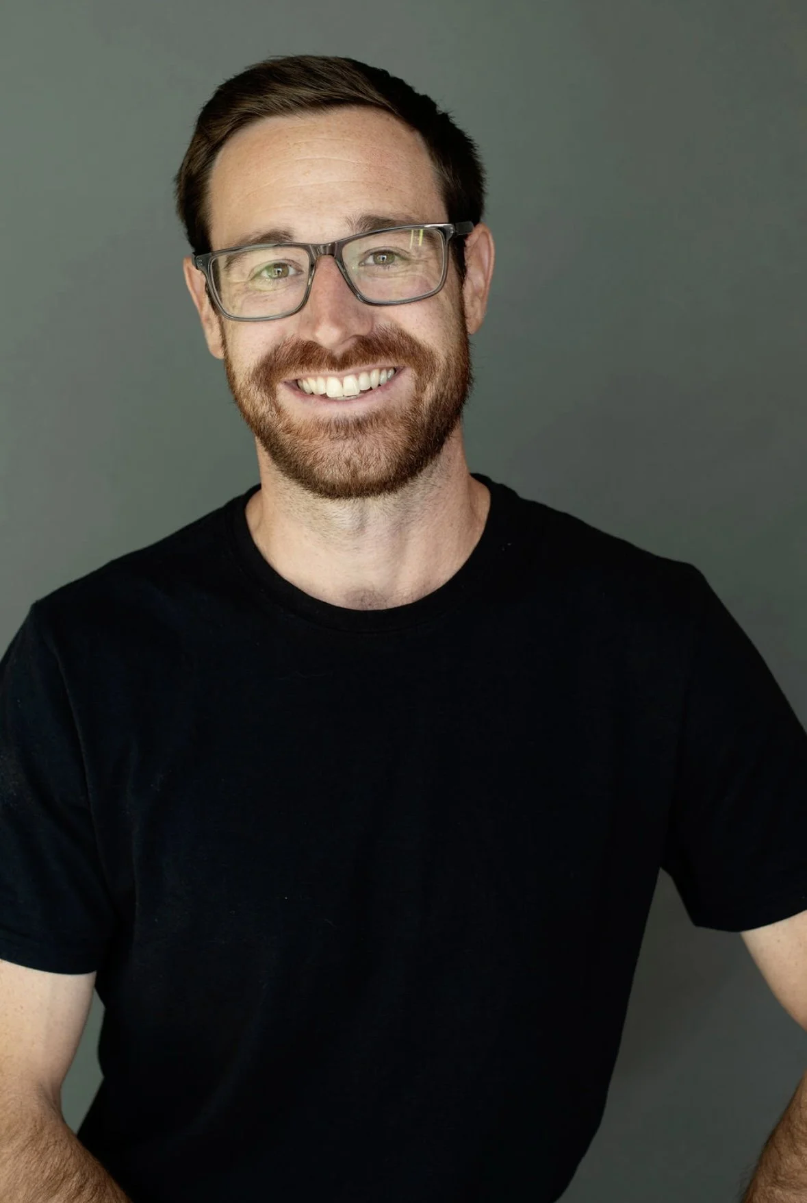 A smiling man with glasses and a beard, wearing a black t-shirt, against a grey background.