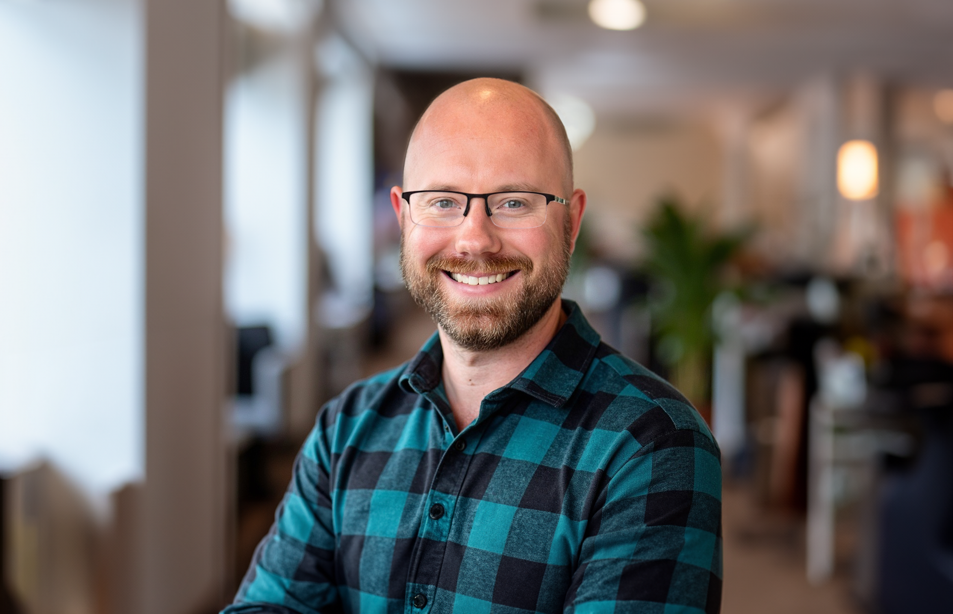 A smiling man with glasses and a beard wearing a blue plaid shirt, standing in a bright, modern indoor space.