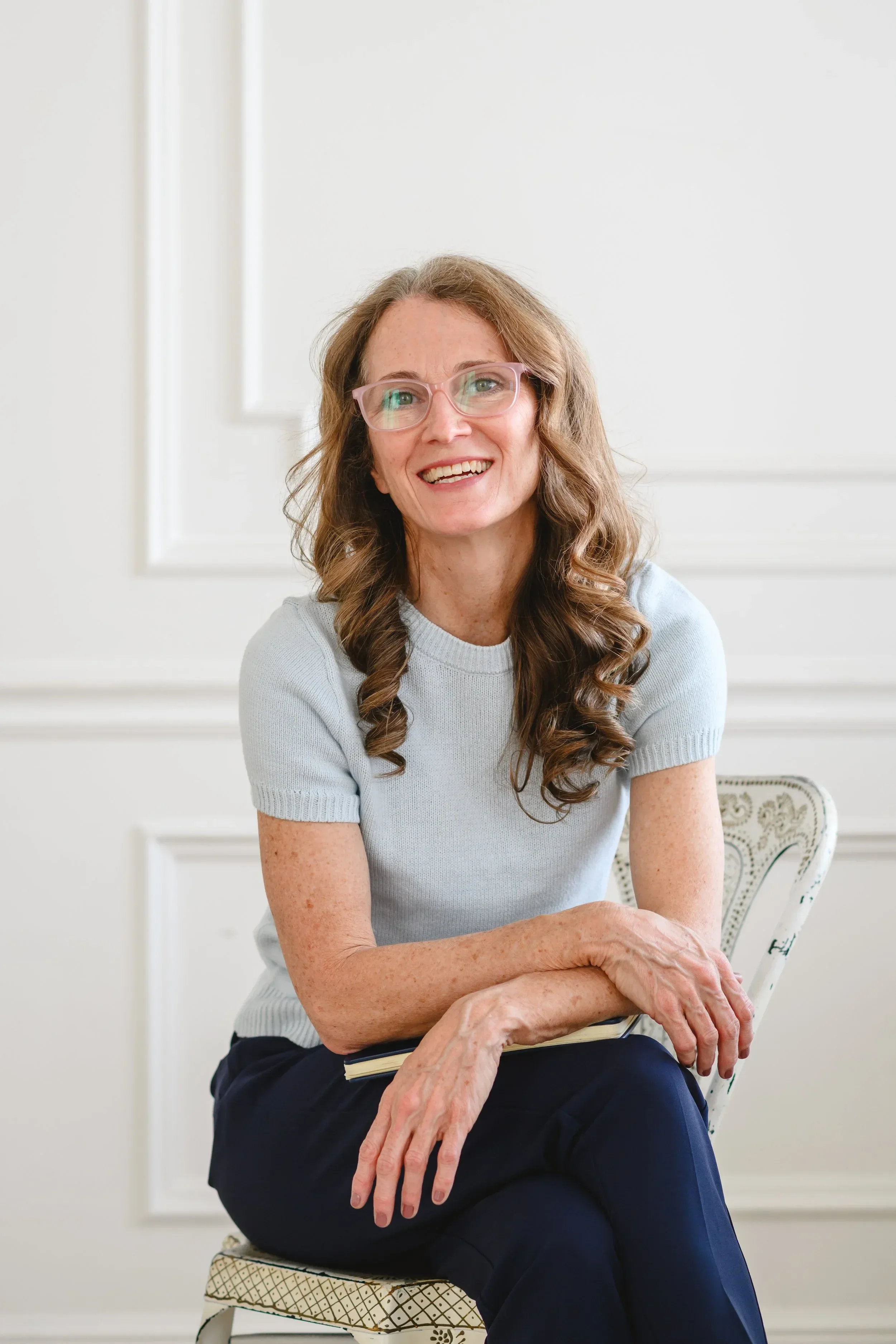 Dr. Servoss wearing glasses sits on a chair, smiling warmly. She wears a light blue sweater and dark pants.