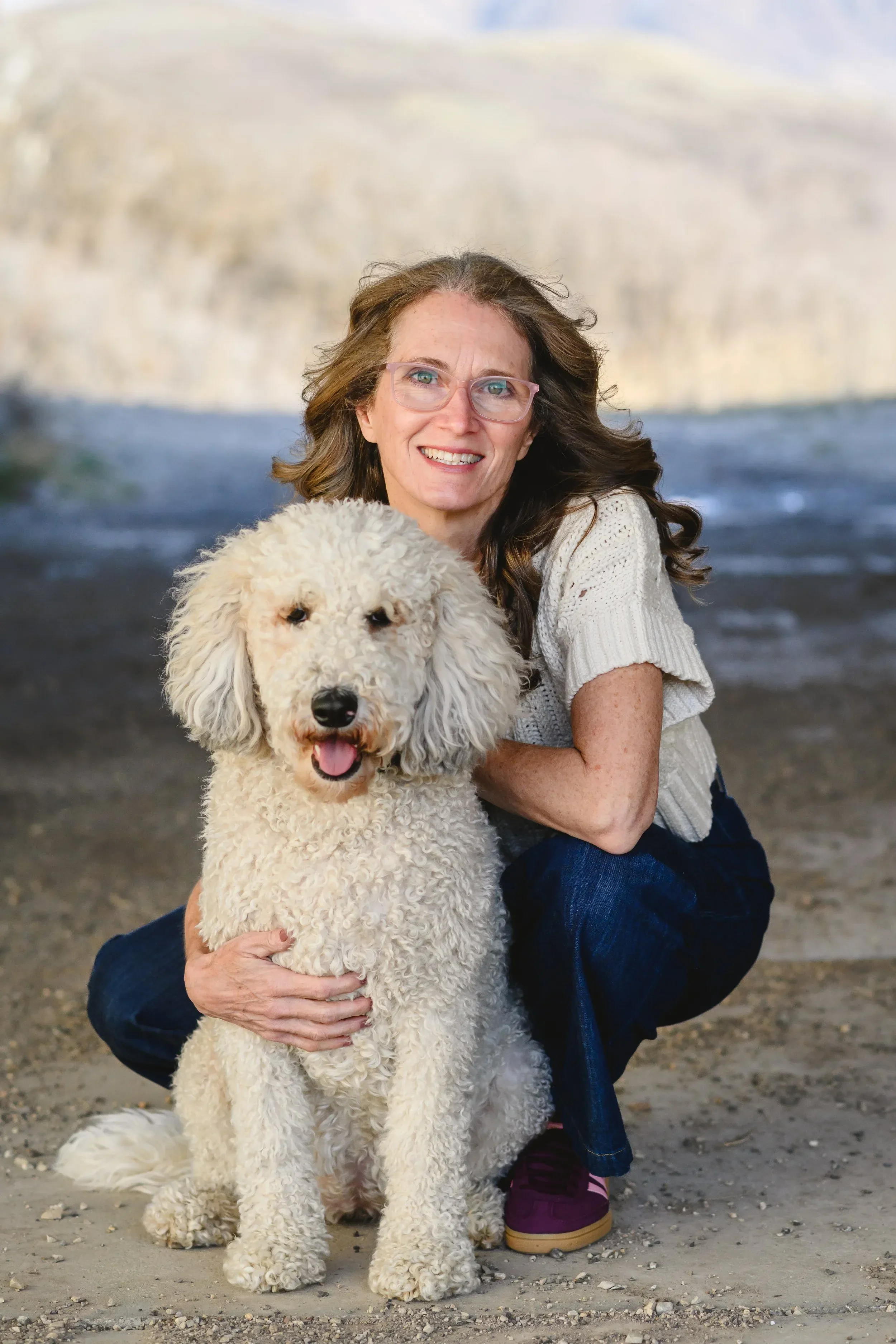 Dr. Servoss kneels beside a fluffy white dog, both smiling. They are outdoors on a dirt path, conveying warmth and happiness.