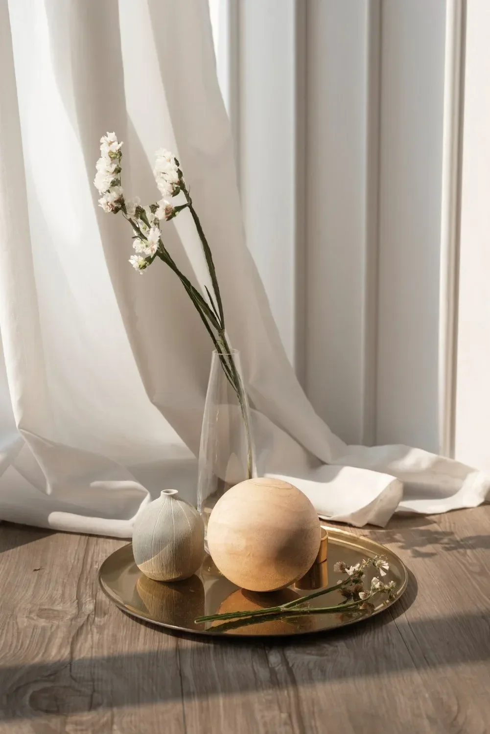 Cozy still life with a glass vase holding white flowers, a wooden sphere, and a small textured ball on a golden tray. Draped curtain in sunlight.