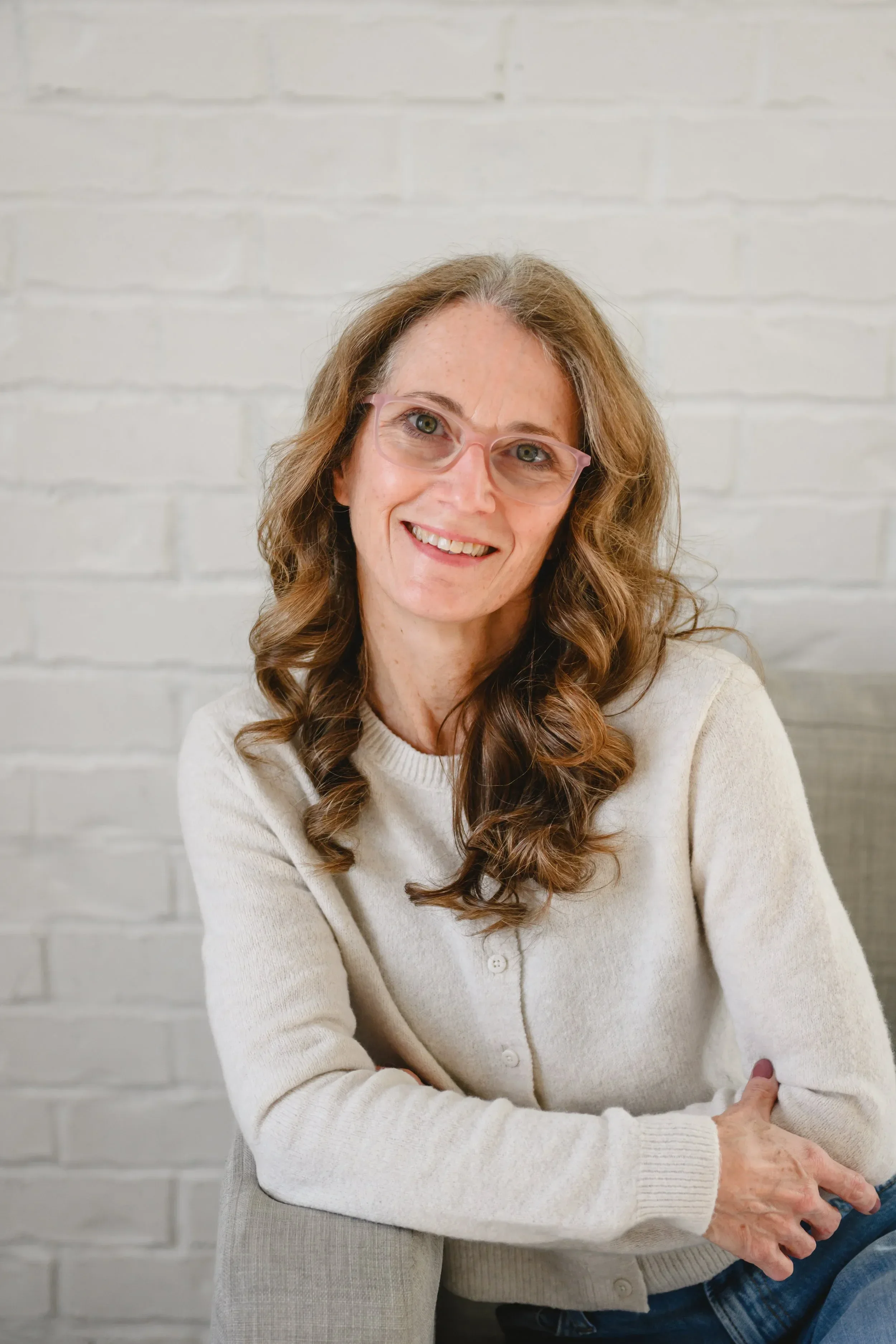 Dr. Servoss sits on a gray chair against a brick wall, wearing a light sweater and blue jeans, conveying warmth.