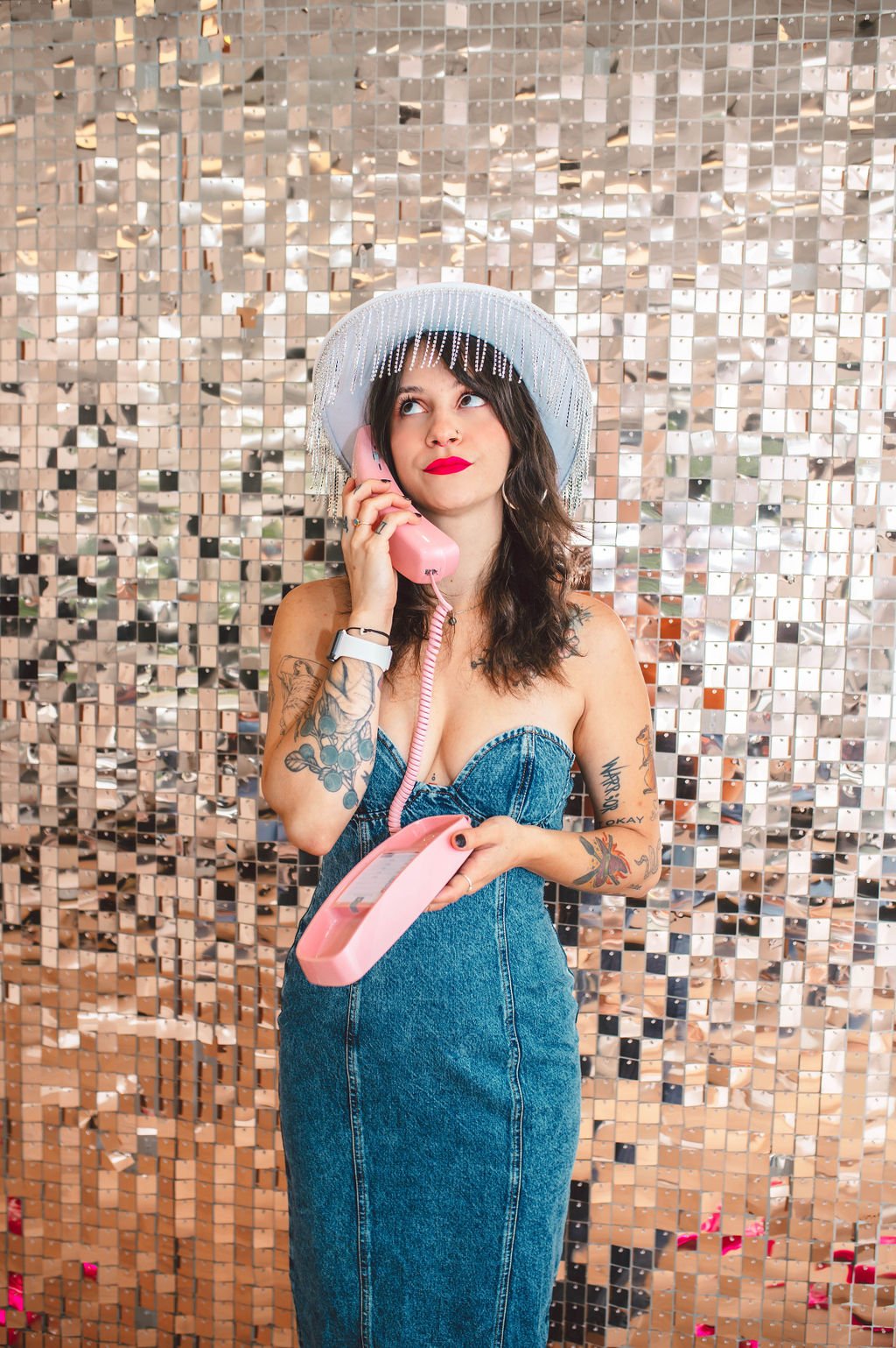 A woman with tattoos wearing a wide-brimmed hat with fringes, holding a pink vintage rotary phone to her ear, standing in front of a reflective mosaic wall.