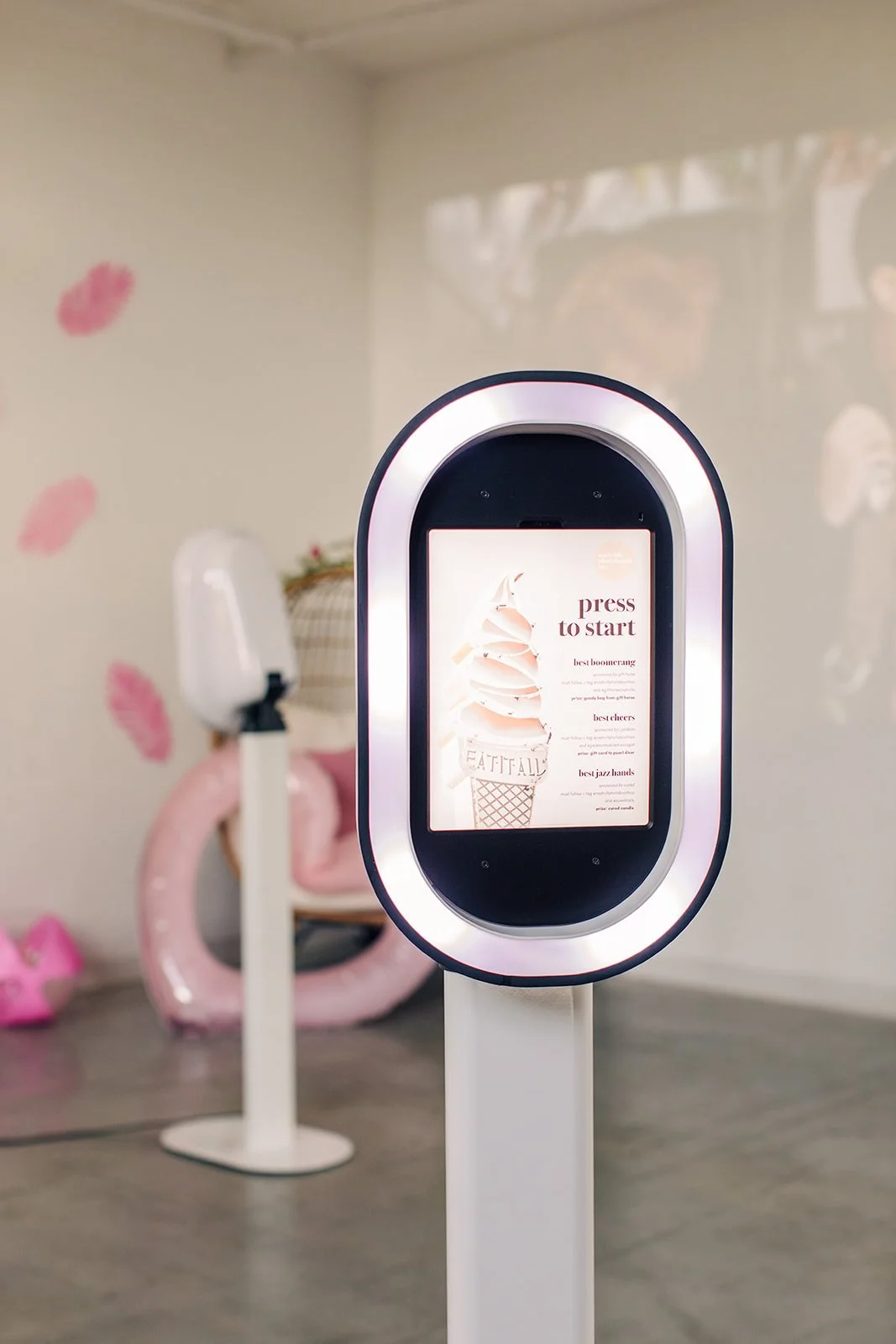 Kiosk with digital screen showing instructions to press to start, featuring an ice cream cone graphic, in a decorated indoor space with pink decorative items in the background.