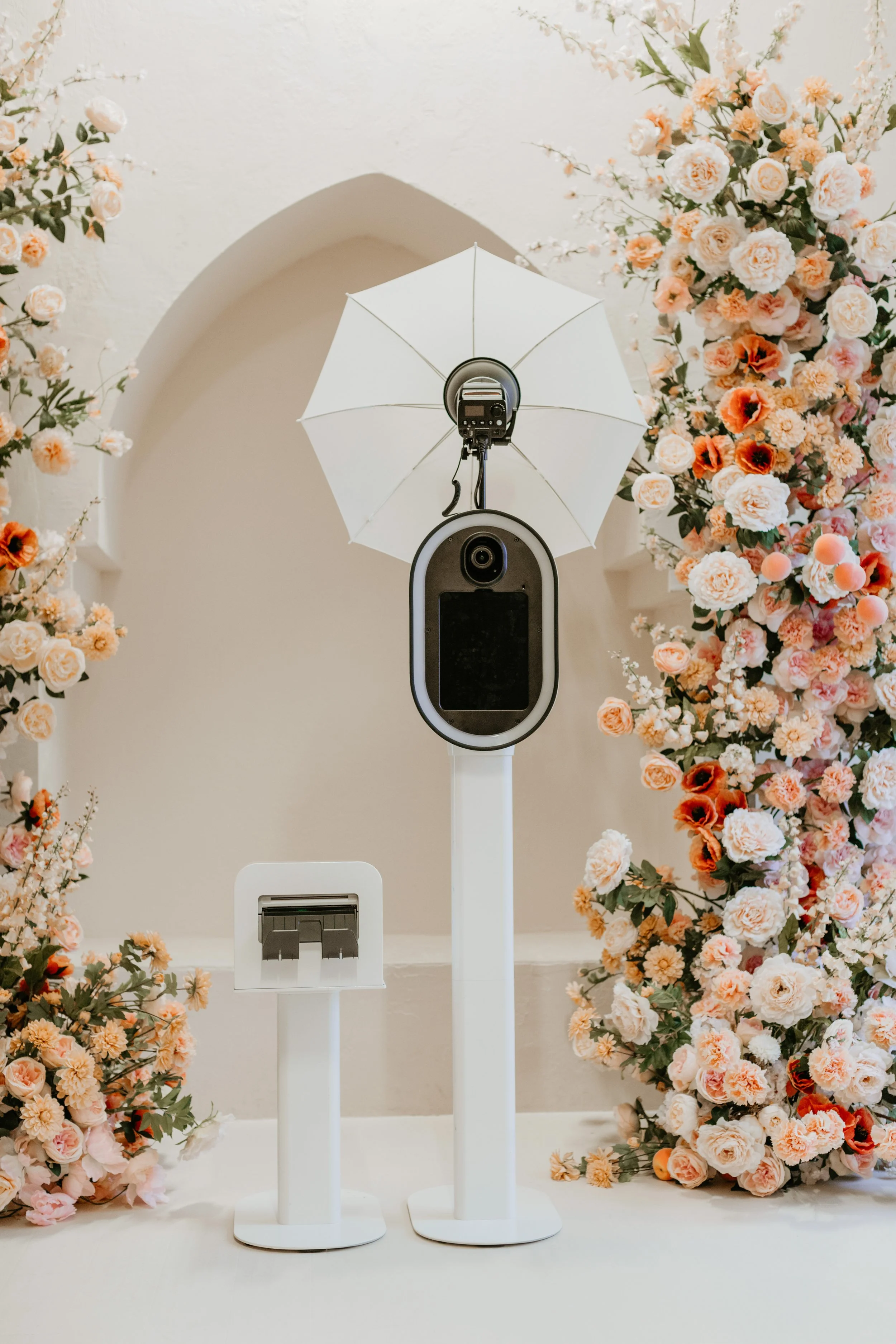 Photobooth setup with a large floral arrangement, a screen and camera, enclosed in a standing frame, with a white umbrella for lighting, at a wedding or event venue.