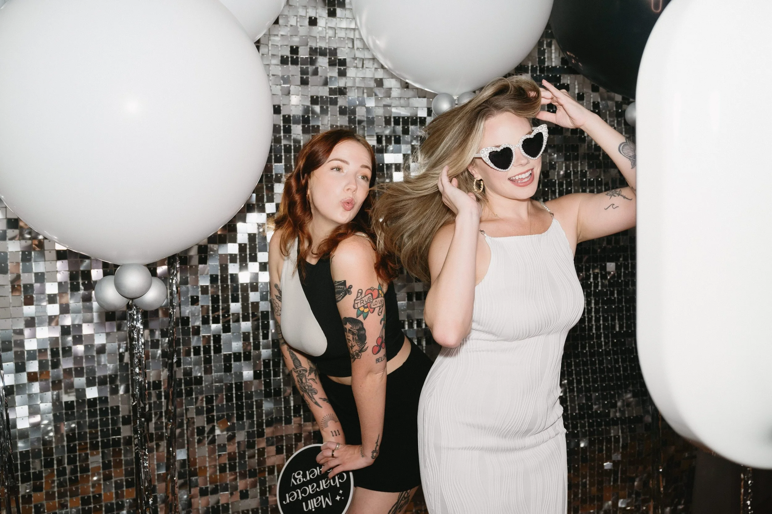 Two women at a celebration with black, white, and silver balloons and a sparkling backdrop. One woman is wearing heart-shaped sunglasses and a white dress, and the other woman is in a black and white dress, making a kissing pose.